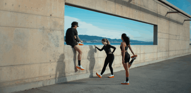 Three young women outdoors in sportswear near a concrete wall with a rectangular window, overlooking a body of water and mountains, with one sitting on the wall and two standing, all engaging in conversation.
