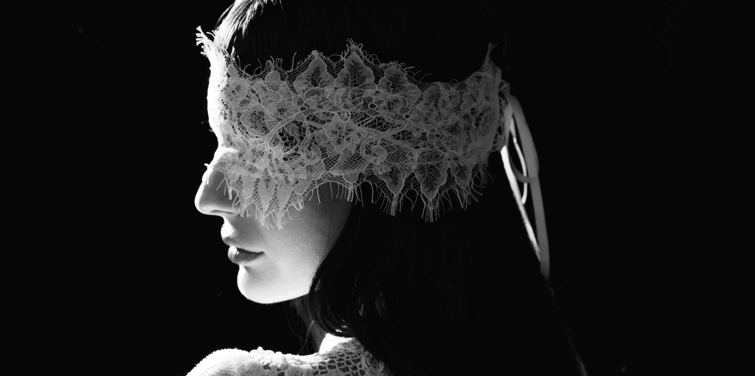 A woman in profile with a lace eye mask and dark hair, illuminated by a soft light against a black background.