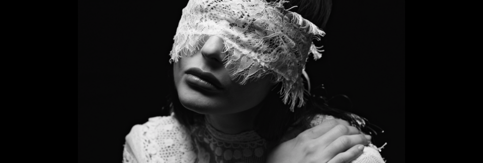 A woman with lace blindfold covering her eyes, dark lipstick, and vintage clothing posing with her hand on her shoulder in a black and white photo.