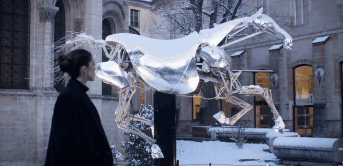 A person standing outdoors in winter, observing a large, metallic horse sculpture with translucent wings on a city street.