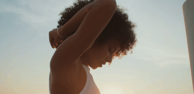 A young woman with curly hair standing outdoors during sunset, adjusting her hair with her arms raised.