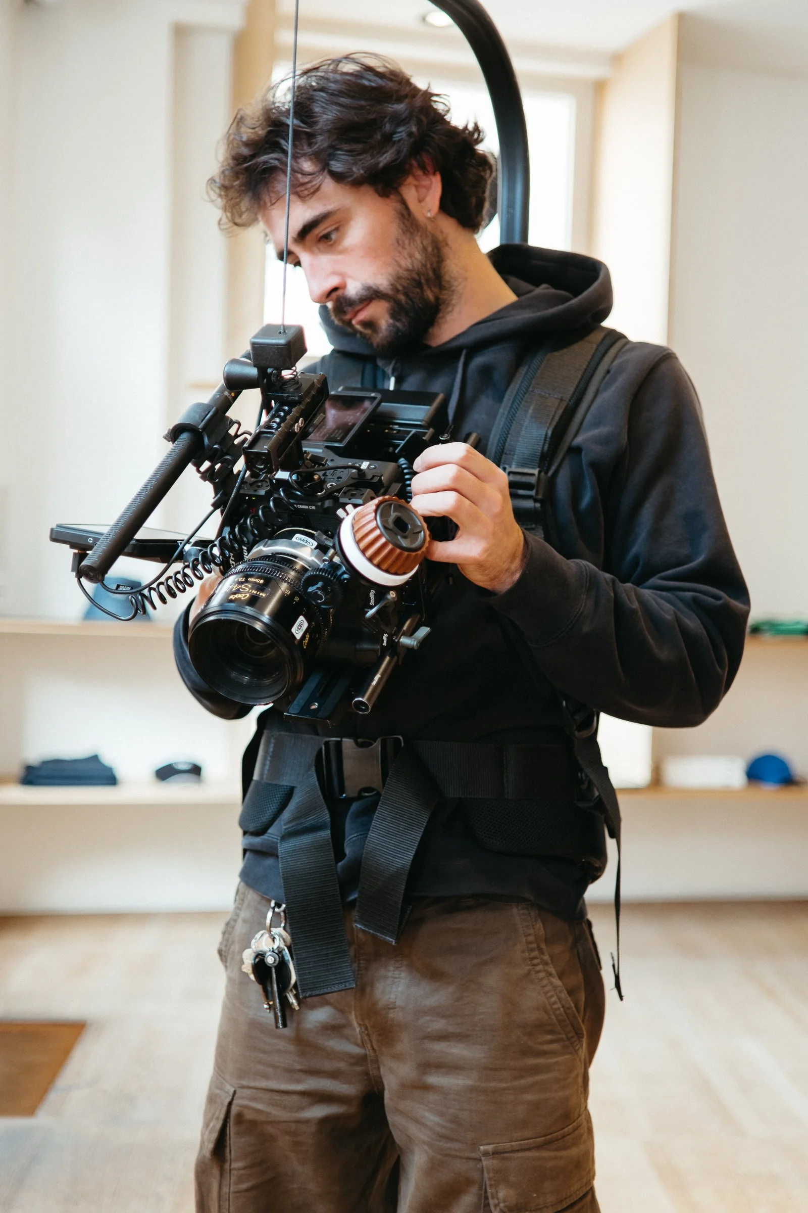 A man with brown cargo pants and a black hoodie, carrying a professional camera rig, is looking down and adjusting the equipment in an indoor setting with beige walls and a window in the background.