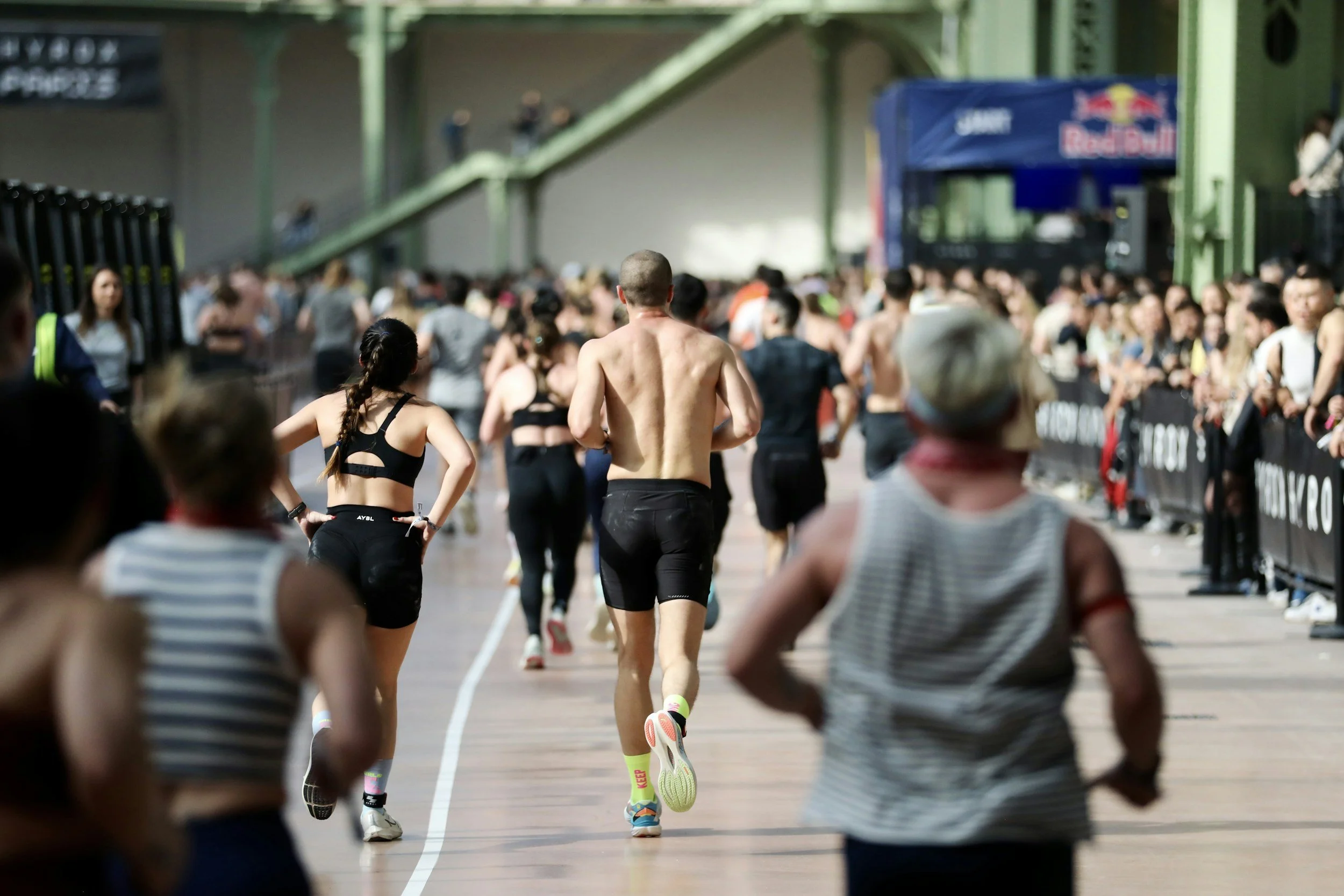 People running in a Hyrox race at an indoor track with a crowd watching and green structural beams in the background.