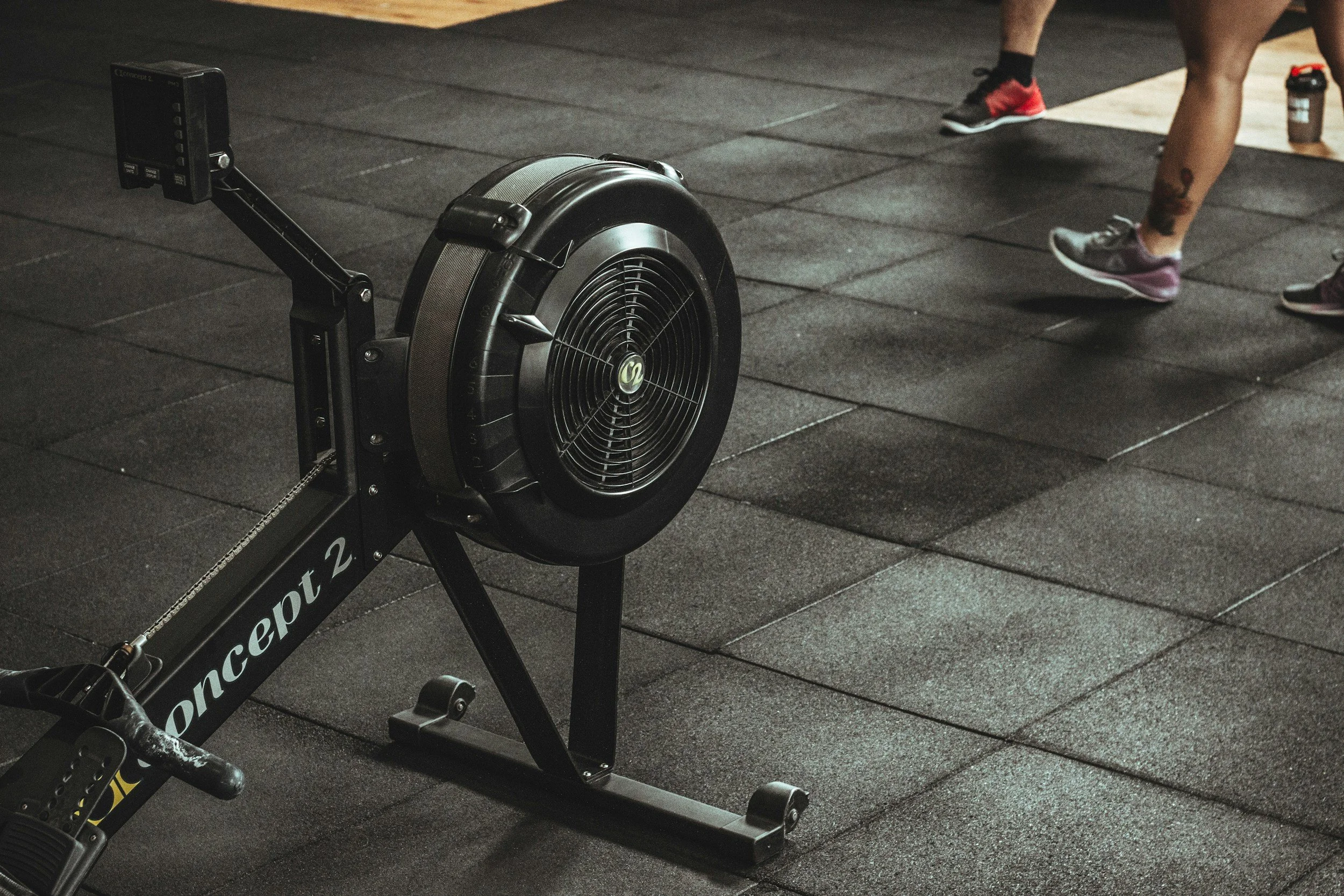 Indoor gym with a seated rowing machine labeled 'Concept 2' and a person wearing pink and gray athletic shoes and black socks, standing on a rubberized workout floor. Hyrox.
