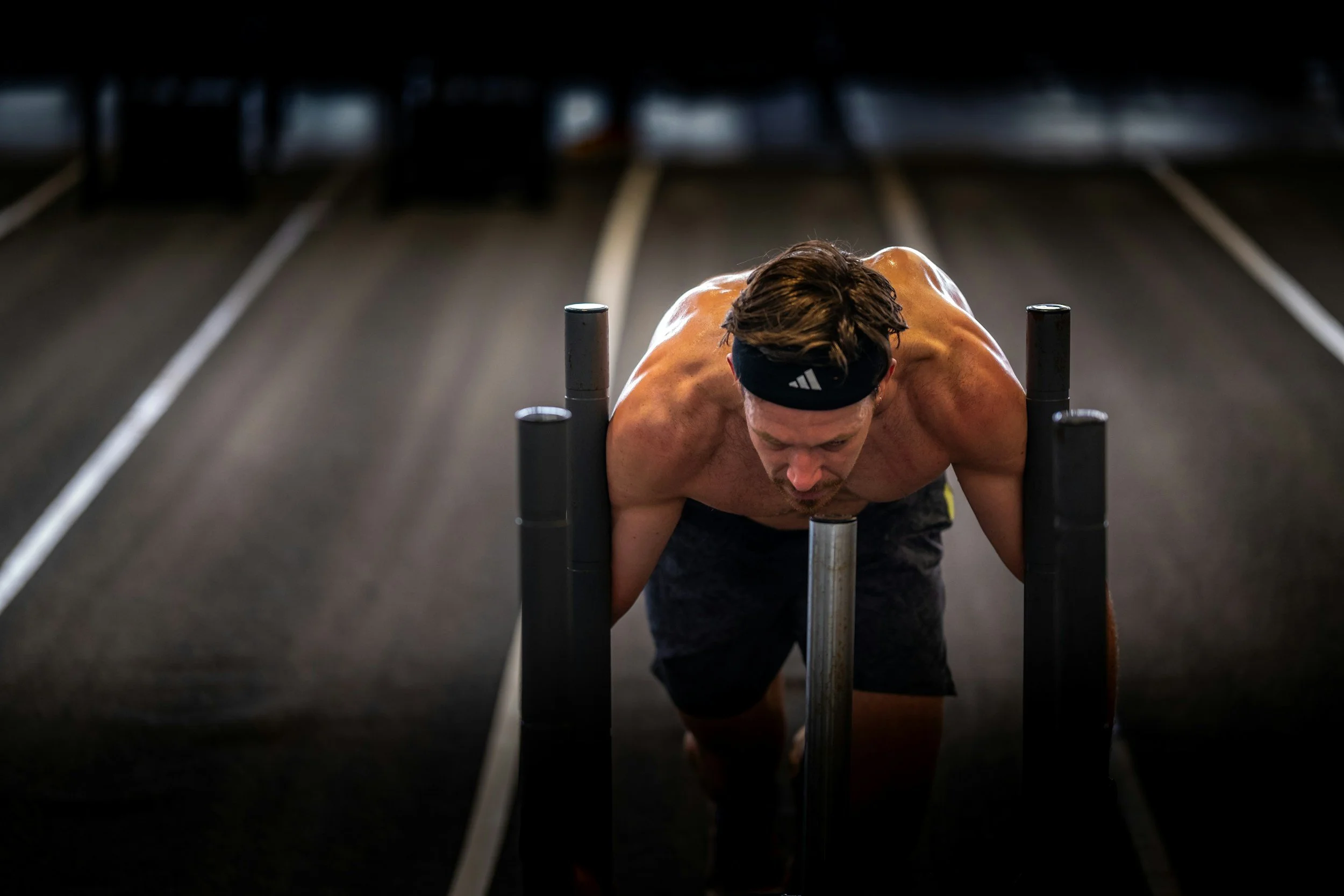 A man in athletic clothing, wearing a black headband, is performing an exercise on a push-up bar in a gym. The background shows a dark workout area with lines on the floor. Hyrox, near me.