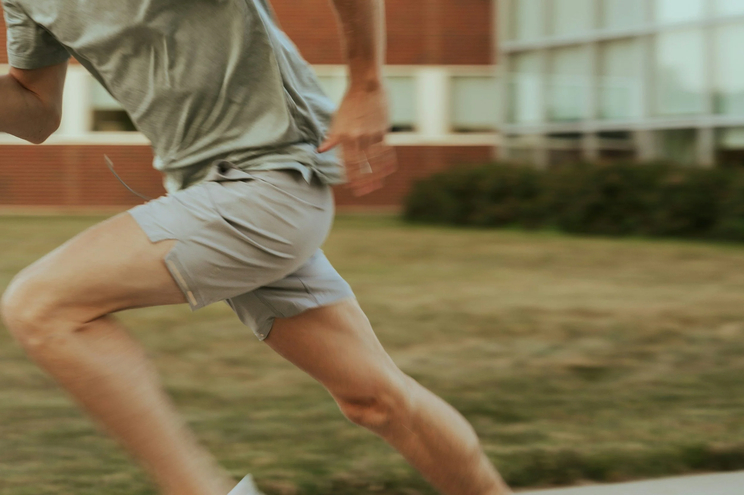 A person running outdoors, wearing a gray t-shirt and beige shorts on a grassy area with a brick building and windows in the background.