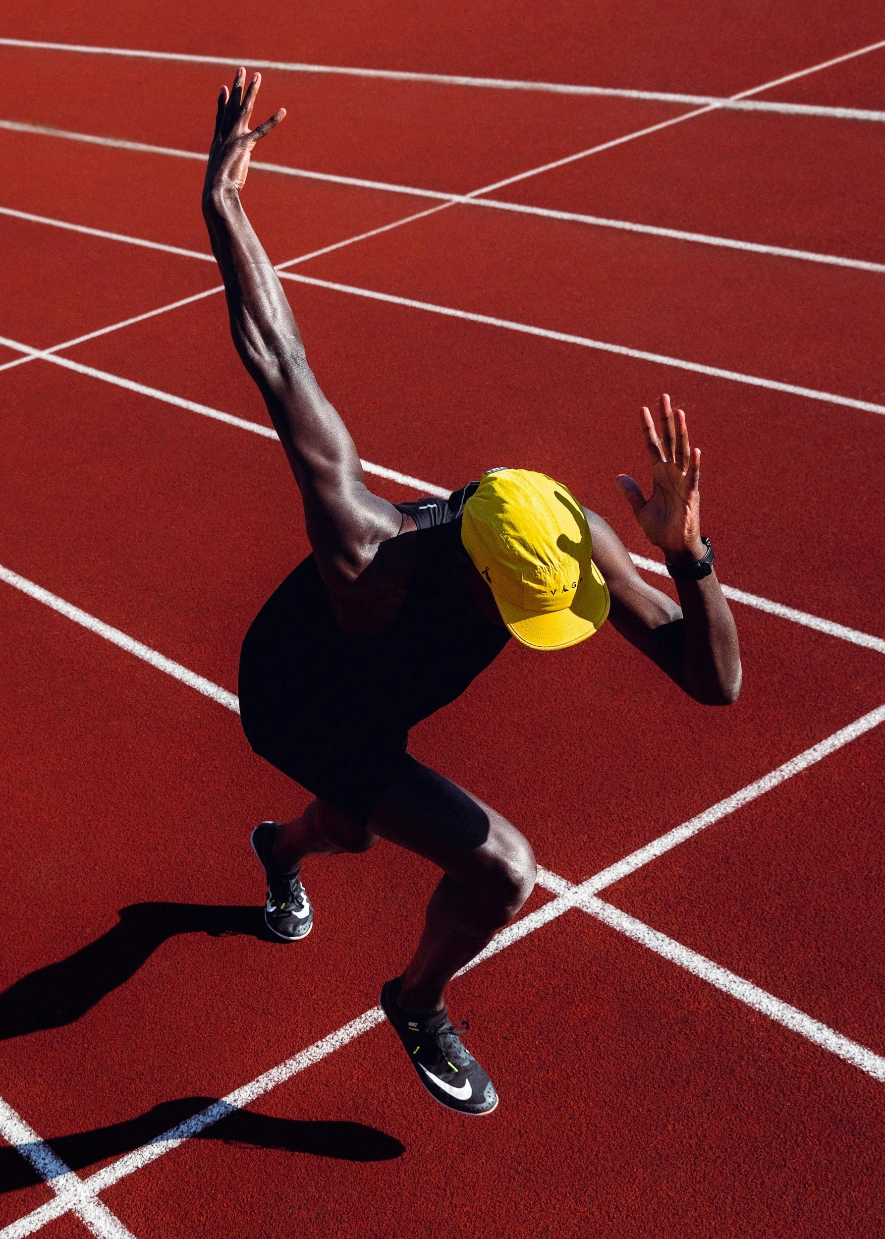 A runner in a yellow cap and black athletic clothing is preparing for a race on a red track, with white lane markings, casting a shadow on the ground. Threshold test, tempo, blood lactate testing.
