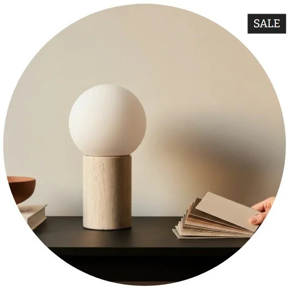 Decorative table lamp with a spherical white shade on a wooden base, with paint color sample cards being held by a hand, and a small stack of books nearby, indicating a sale on home decor items.