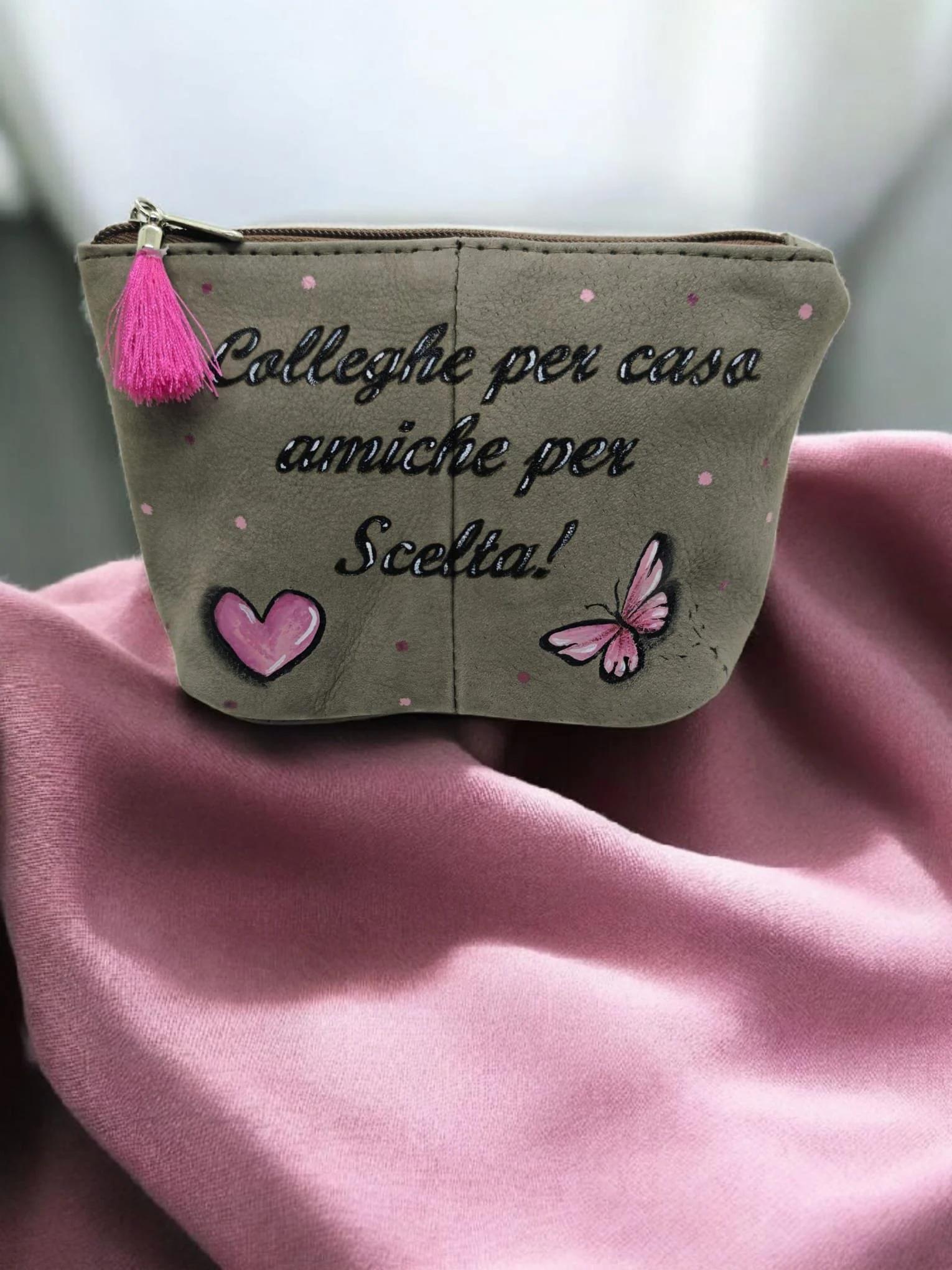 Custodia beige con scritta in nero e decorazioni di farfalle e cuori rosa, con una nappina rosa e una cerniera marrone, posta sopra un tessuto rosa