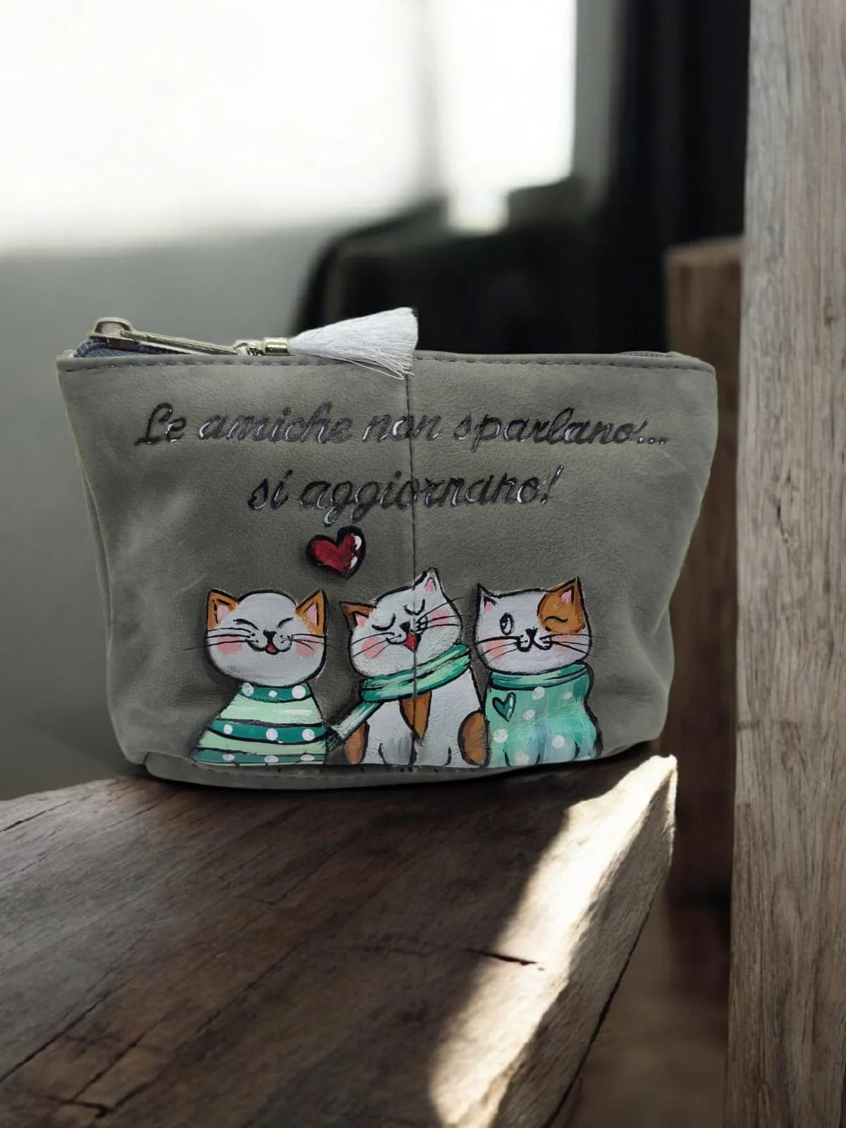 Una pochette di tessuto grigio con ricami di tre gatti stilizzati con sciarpe colorate. Sopra i gatti ci sono le scritte italiane: "Le amiche non sparlano... si aggiustano!" e un cuore rosso.