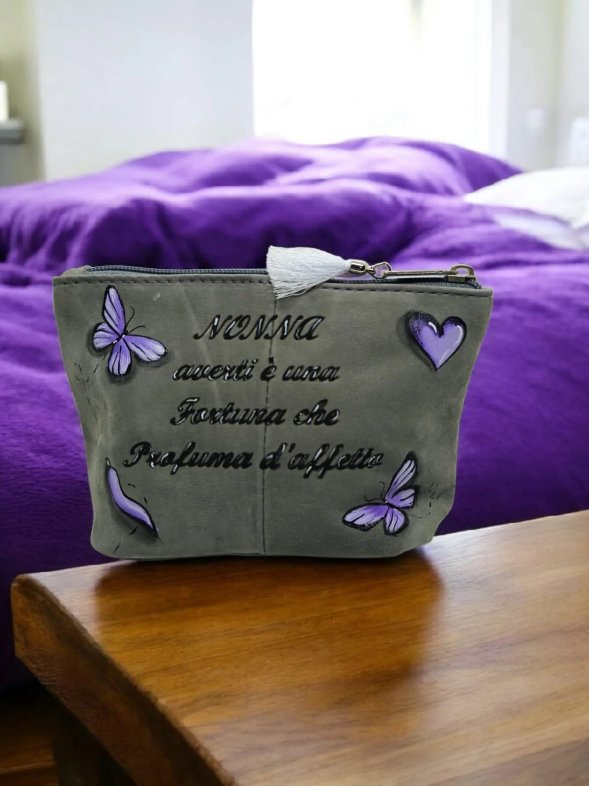 Una borsetta grigia con disegni di farfalle e un cuore viola, appoggiata su un tavolo di legno, con una coperta viola e un letto sopra in background.