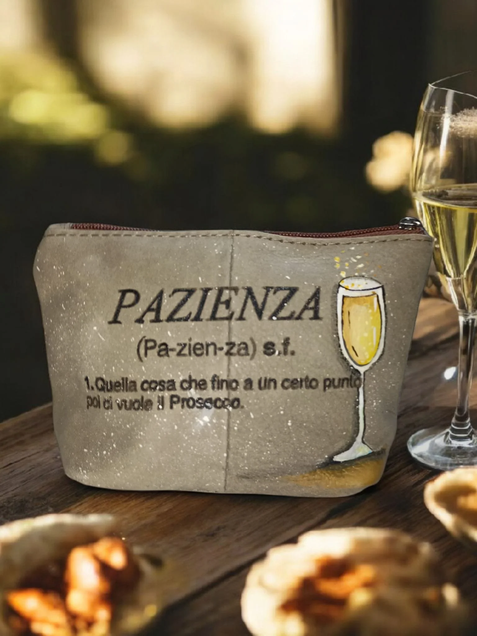 Un sacchetto di paglierino con scritta 'PAZIENZA' e una frase sotto, il tutto accompagnato da un bicchiere di champagne e altri oggetti sulla tavola.