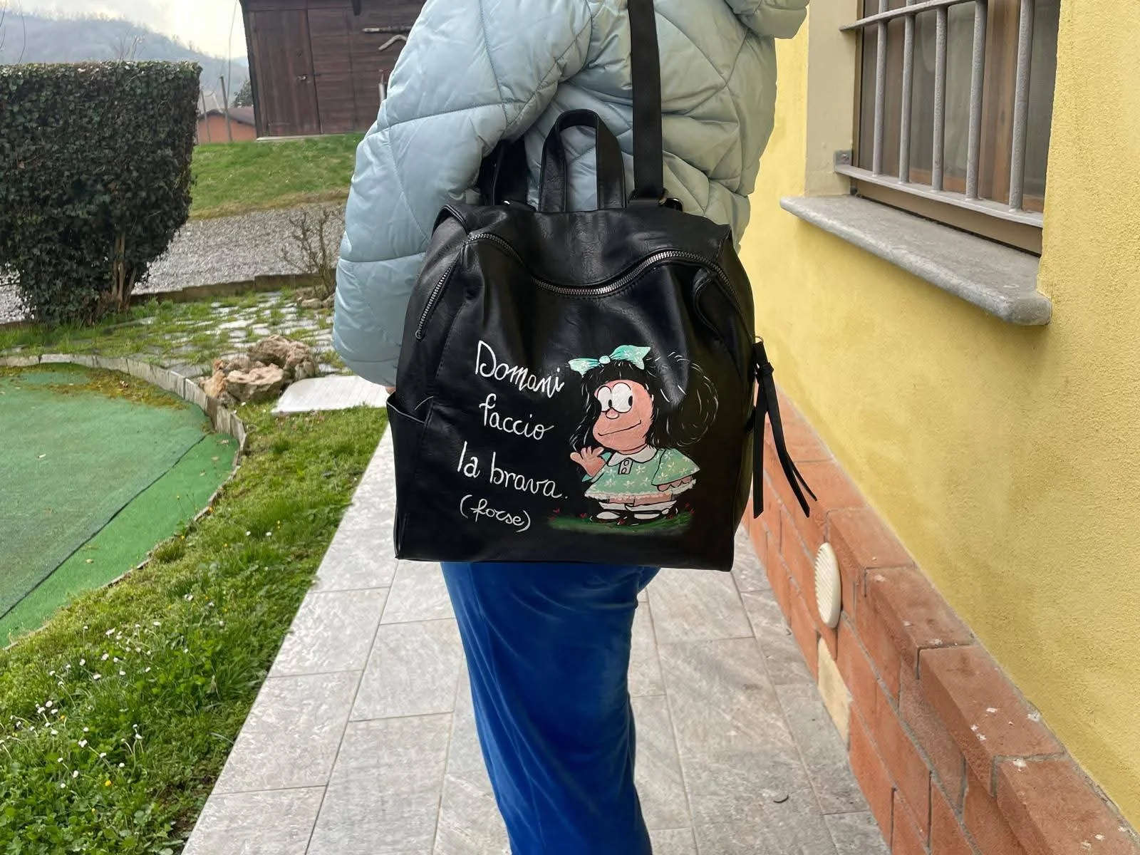 Una persona cammina all'aperto con una borsa di pelle nera decorata con un disegno e una scritta in italiano. La scritta dice: 'Donnivi faccio la brava.' La persona indossa una giacca azzurra e pantaloni blu, e si trova vicino a una casa con pareti g