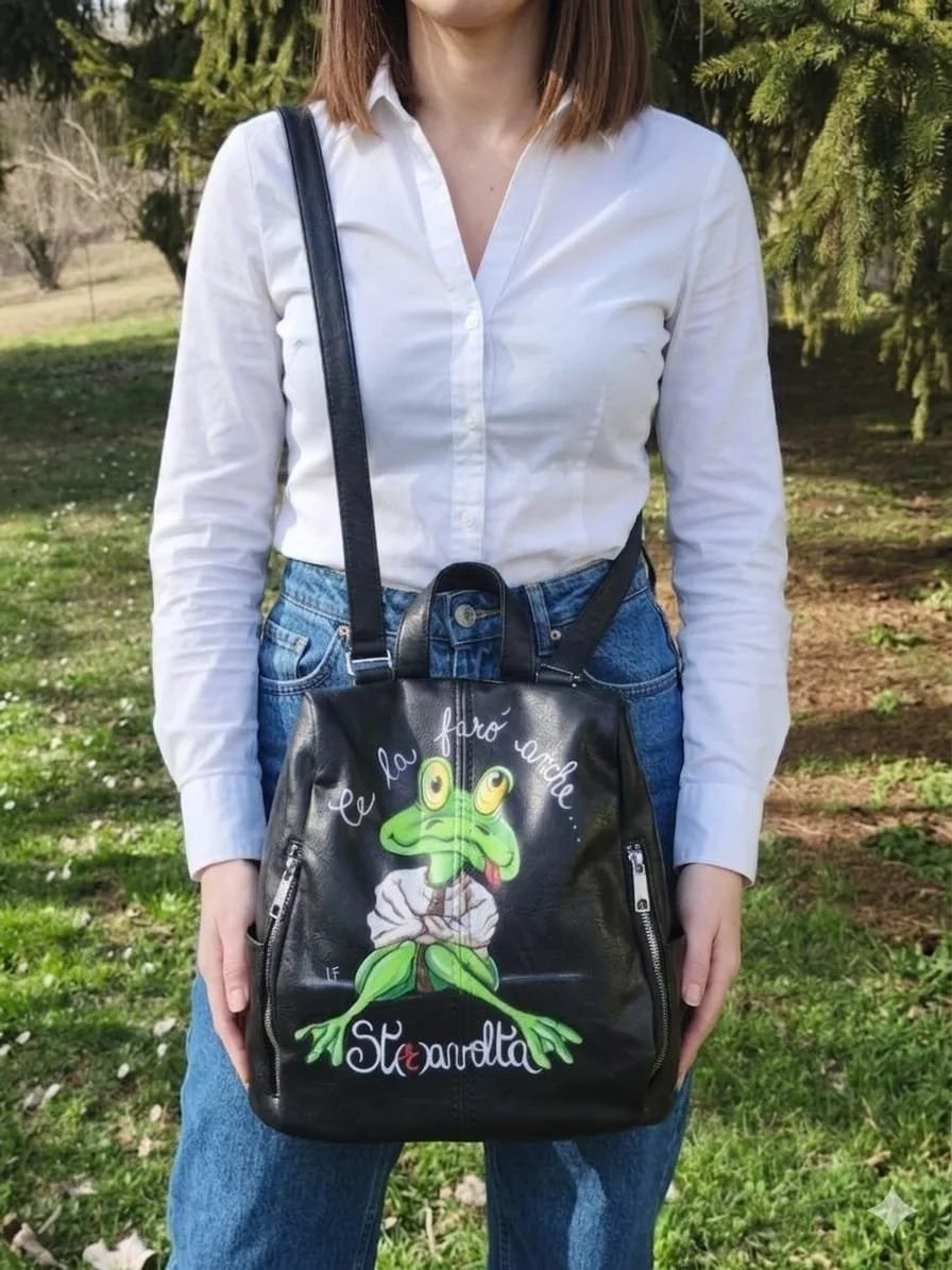 Borsa nera con una stampa di una rana con occhi grandi e testo in italiano che dice 'Ce la farà anche...' e una parola 'Stenardta' in stile artistico, indossata da una persona con camicia bianca e jeans, all'aperto con sfondo di alberi e prato.