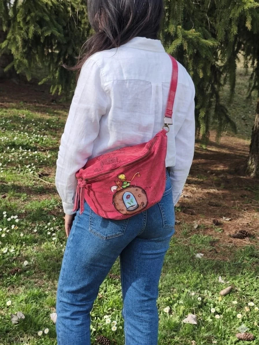 Una persona con camicetta bianca e jeans blu indossa una borsetta rossa con disegni di un personaggio e una pigna, situata sulla schiena, all'esterno tra alberi e prato.