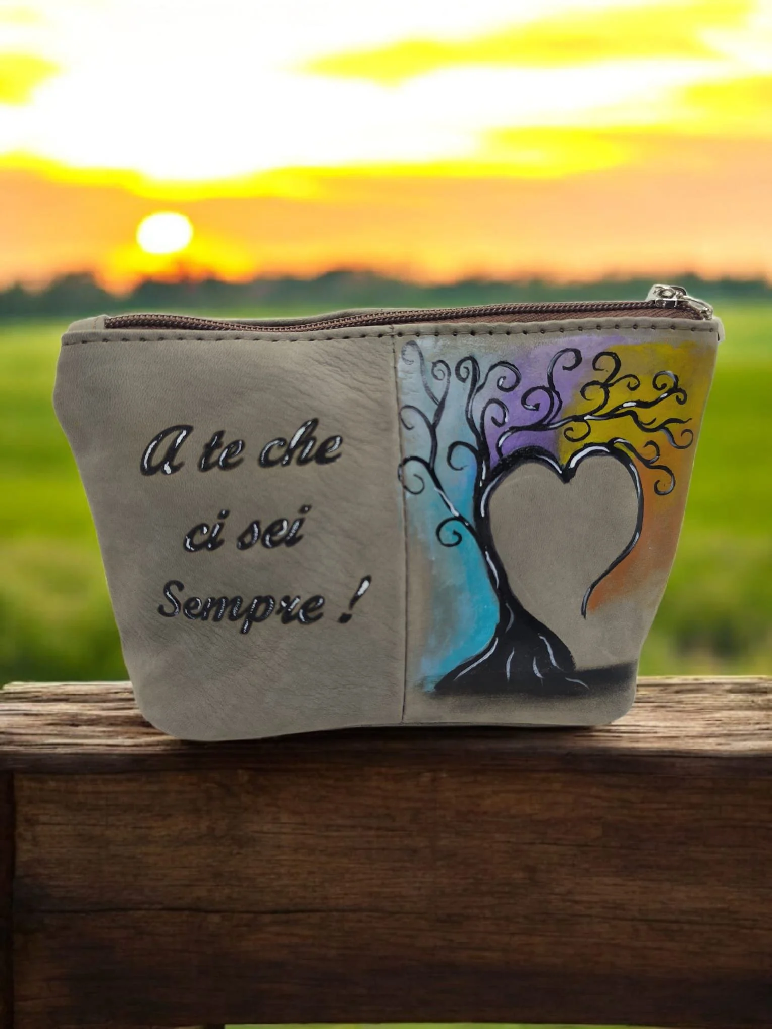 Un sacco colorato con un disegno di un albero stilizzato con cuore e scritte in italiano, posato su una superficie di legno con un tramonto sullo sfondo.