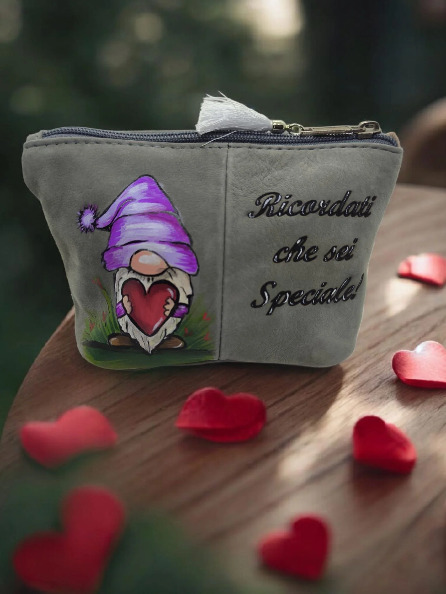 Una piccola borsa di stoffa grigia con una zip, decorata con un disegno di un gnomo con cappello viola, e scritto in italiano "Ricordati che sei speciale!". Circondata da petali di cuore rossi.
