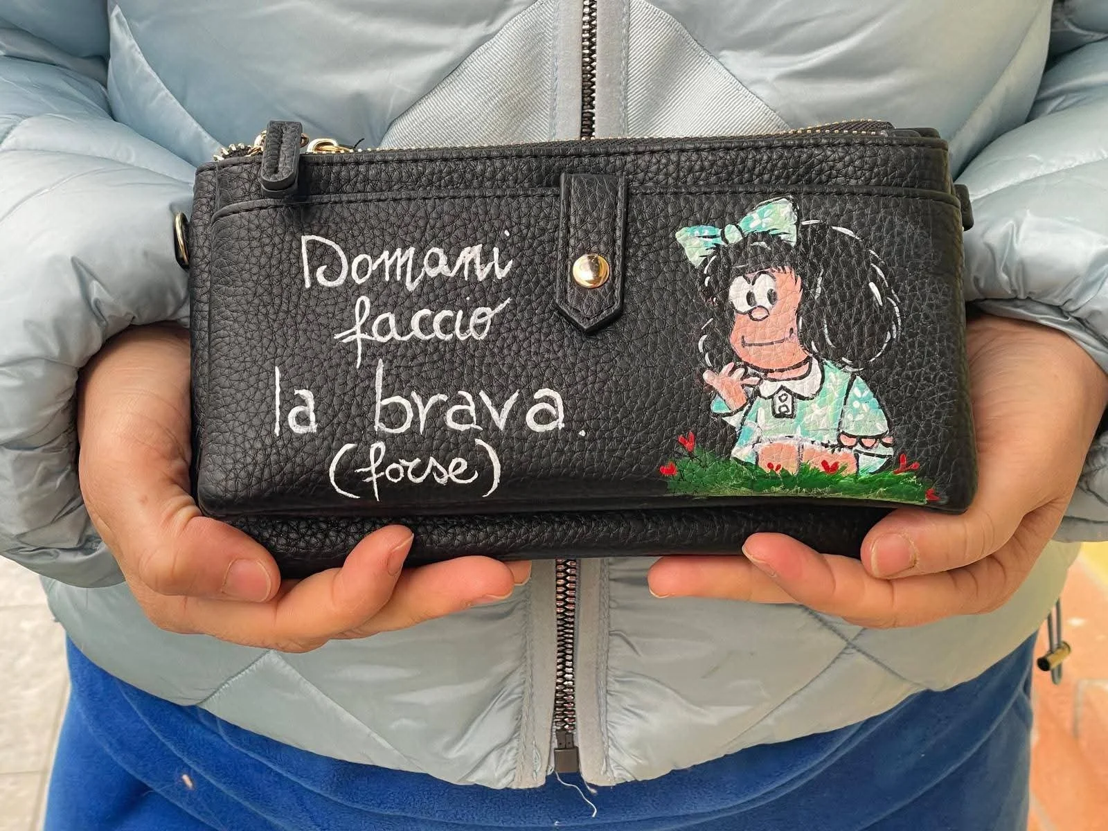 Una persona sta tenendo una borsetta di pelle nera con scritte bianche e un disegno di una bambina stilizzata con capelli ricci e capelli nero con una fascia verde, circondata da dei fiori rossi. Sulla borsa c'è una frase in italiano: 'Domani faccio 