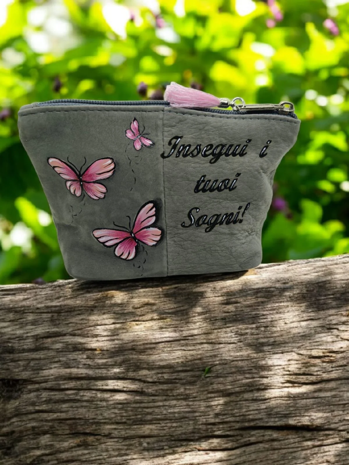 Una pochette grigia decorata con farfalle rosa e il testo 'Inseguì i tuoi Sogni!' scritto sopra. La pochette è appoggiata su un tronco di legno, con uno sfondo di foglie verdi.