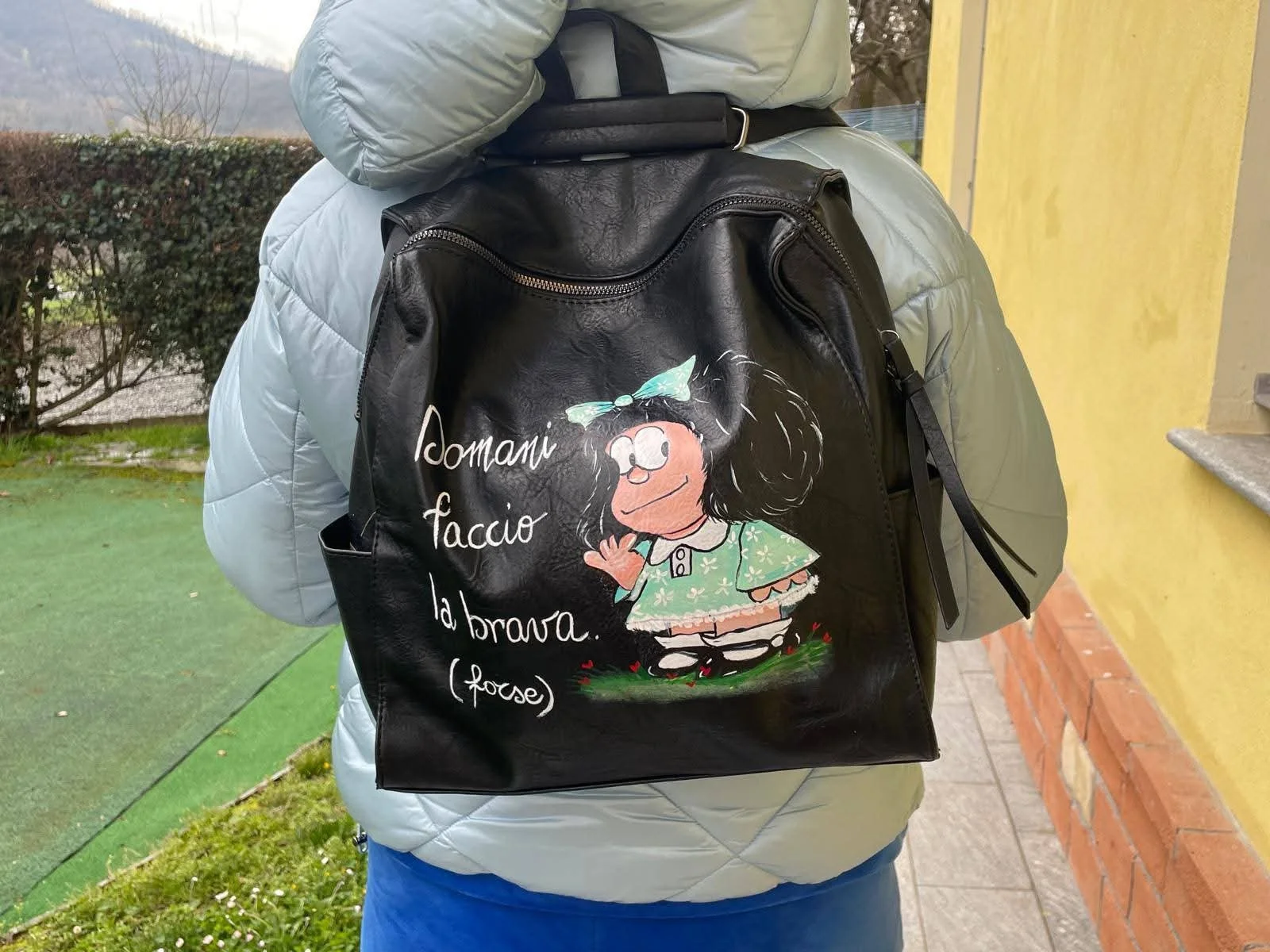 Zaino nero con disegno di una bambina con capelli ricci e fiocco azzurro, con scritta in italiano 'Idomani faccio la brava (poche)'. Indossato da una persona con giacca grigia e pantaloni blu, all'aperto vicino a un muro giallo e un'area verde.