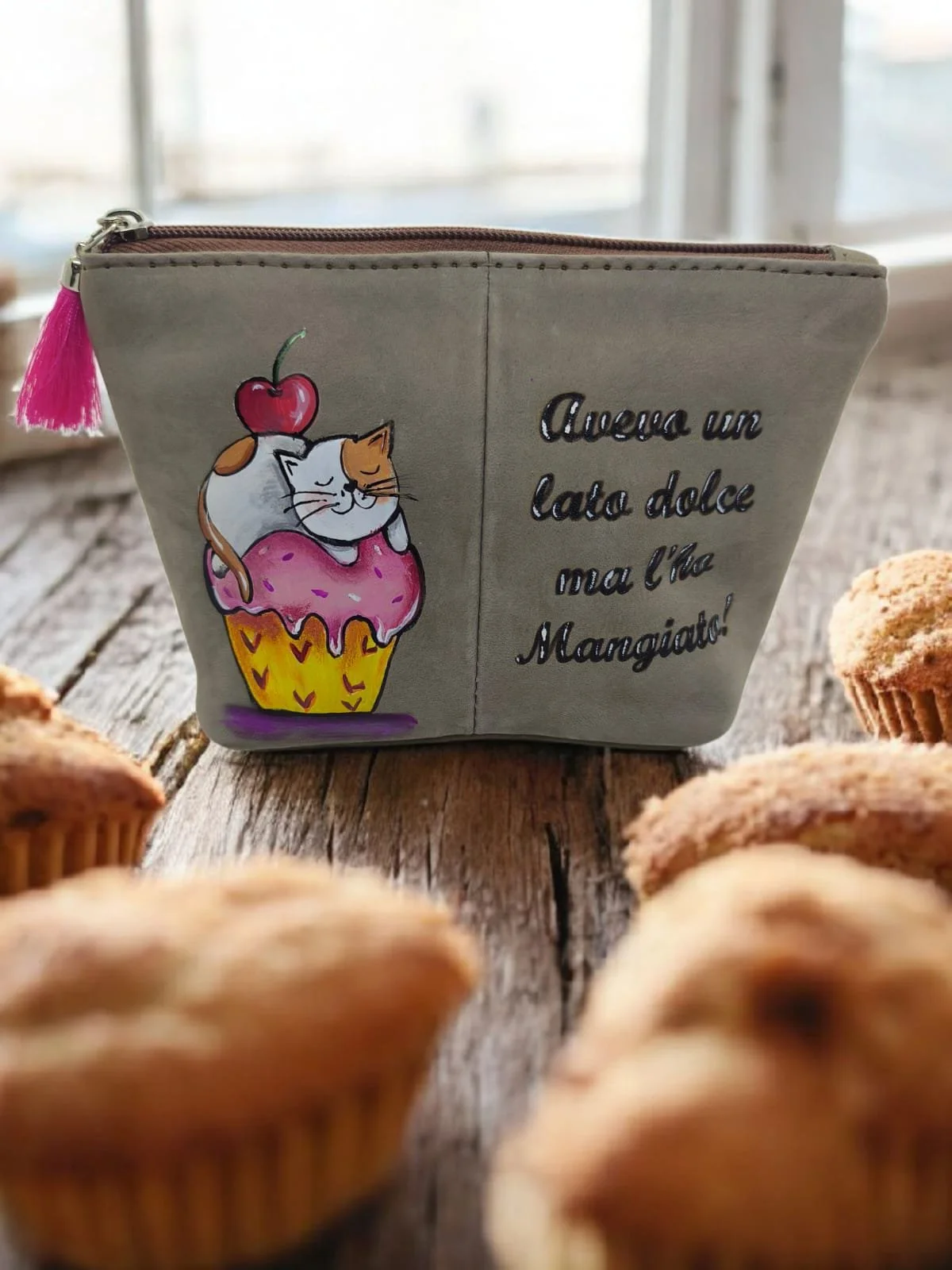 Borsa con illustrazione di un gatto, una ciliegia e una coppetta di gelato, con scritta in italiano. Davanti a una finestra, su un tavolo di legno con muffin sparsi.