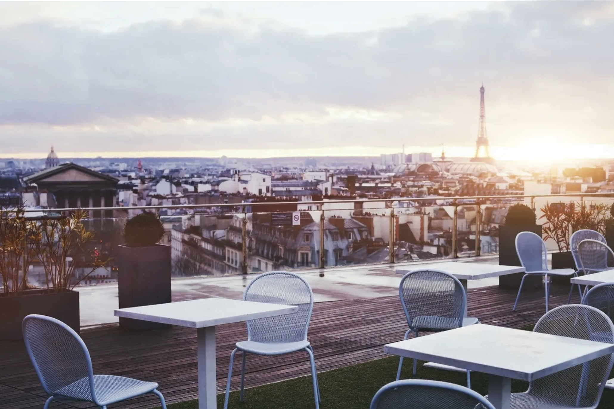 Rooftop terrace with white tables and chairs overlooking the Paris skyline, including the Eiffel Tower at sunset with cloudy sky.