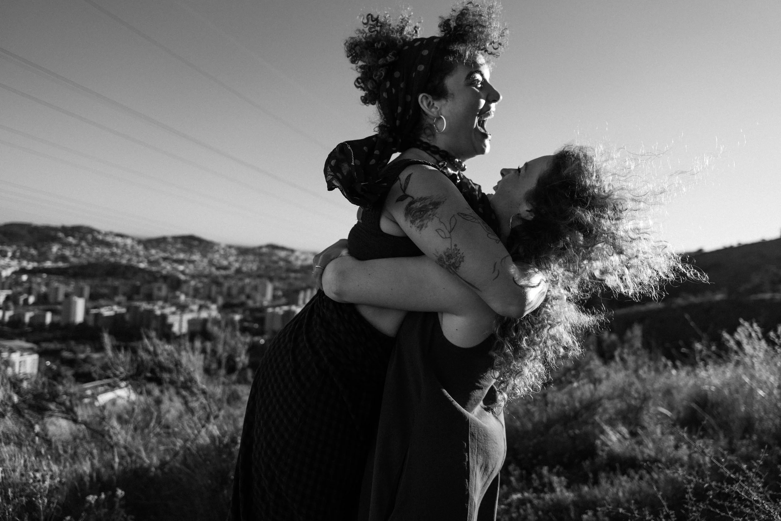 Two women embrace outdoors with hills and a city skyline in the background, one woman lifting the other, both smiling, black and white photo.