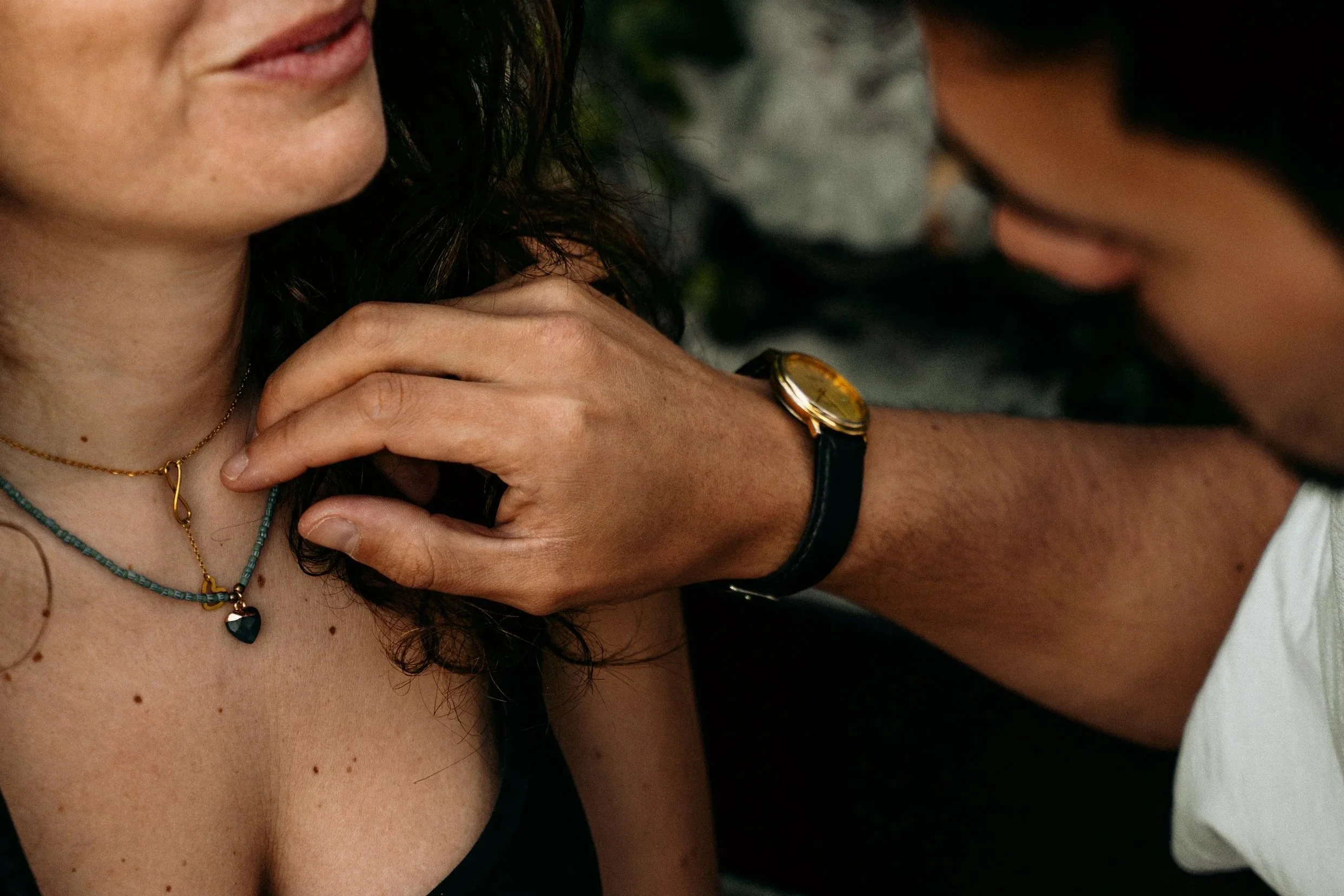 Close-up of a woman with dark hair, wearing necklaces, and a gold watch on her wrist, holding her hand on her shoulder, with a man's hand gently resting on her shoulder.