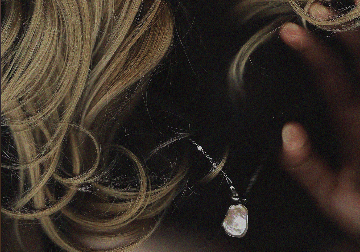 Close-up of blonde curly hair and a hand holding a necklace with a large irregular pearl pendant.