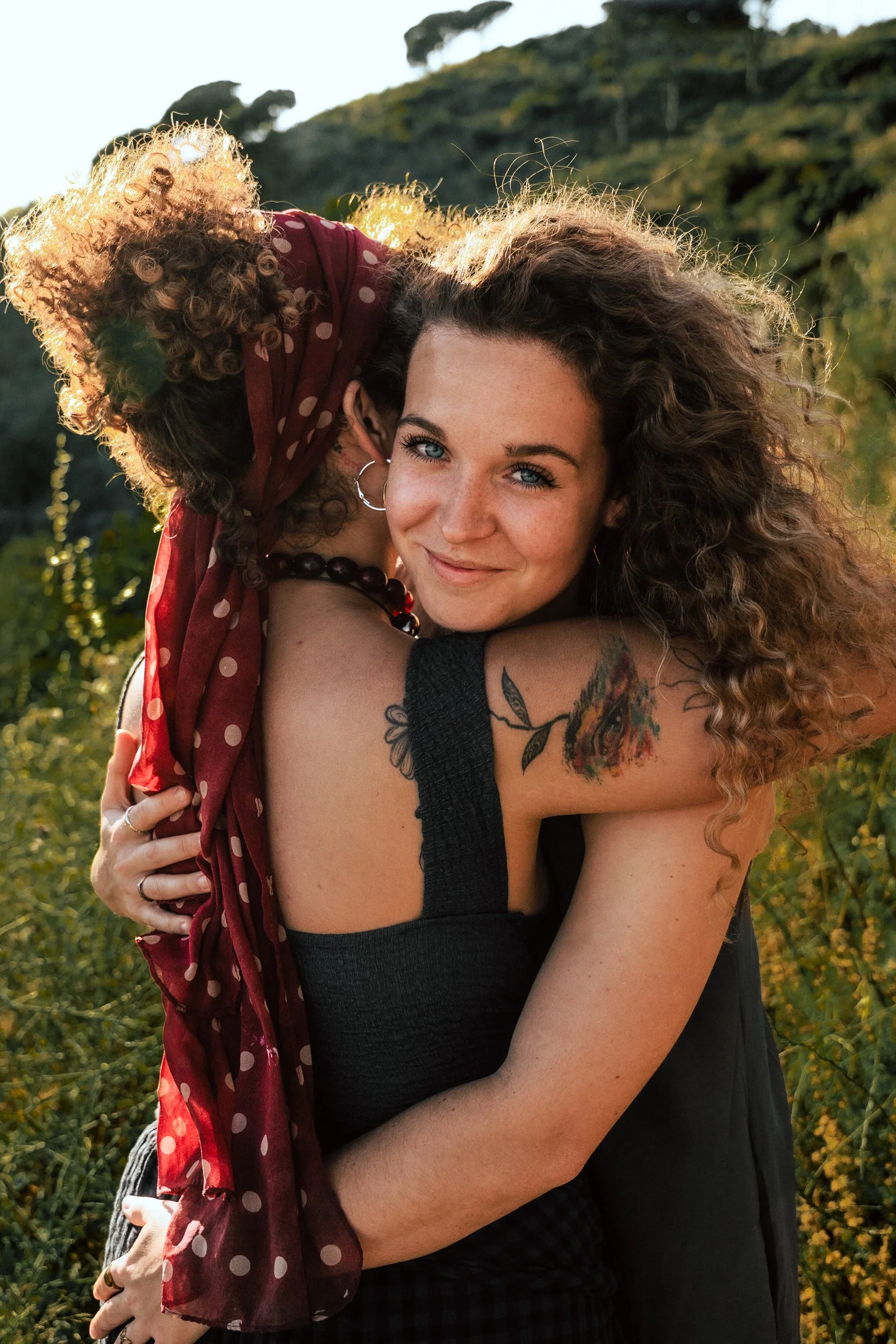 Two women embracing outdoors with a grassy hill in the background. One woman facing the camera has long curly hair, a tattoo on her shoulder, and is smiling. The other woman, with blonde curly hair and wearing a red polka dot scarf, is hugging her fr