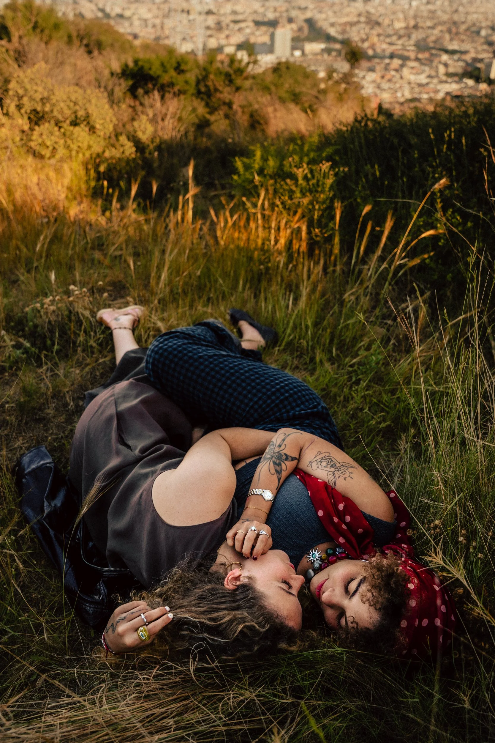 Two women lying on a hillside with Barcelona cityscape behind them — couples portrait session by Whip & Honey
