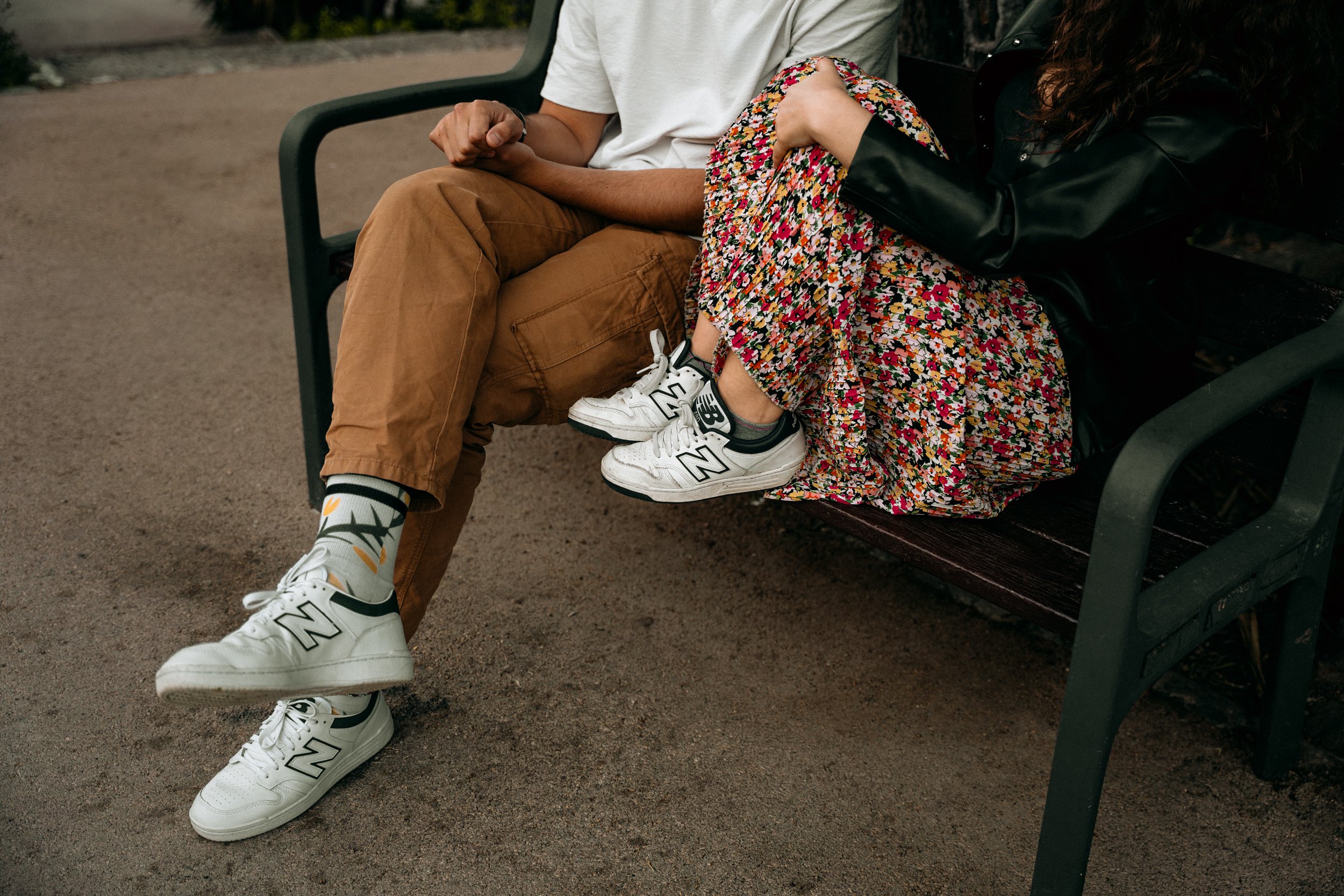 Two people sitting on a park bench, holding hands, with their legs crossed. One person is wearing khaki pants, white sneakers, and floral socks. The other person is wearing a floral skirt and a black leather jacket.