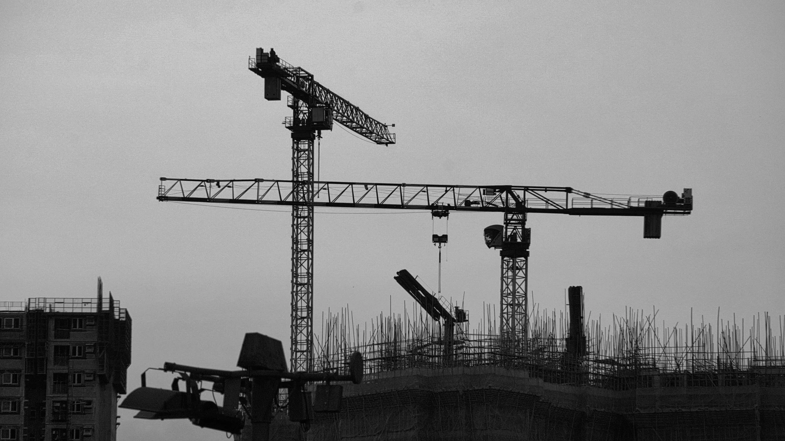 Schwarz-weiß-Bild eines Baukrans auf einer Baustelle, mit Gebäuden im Hintergrund.