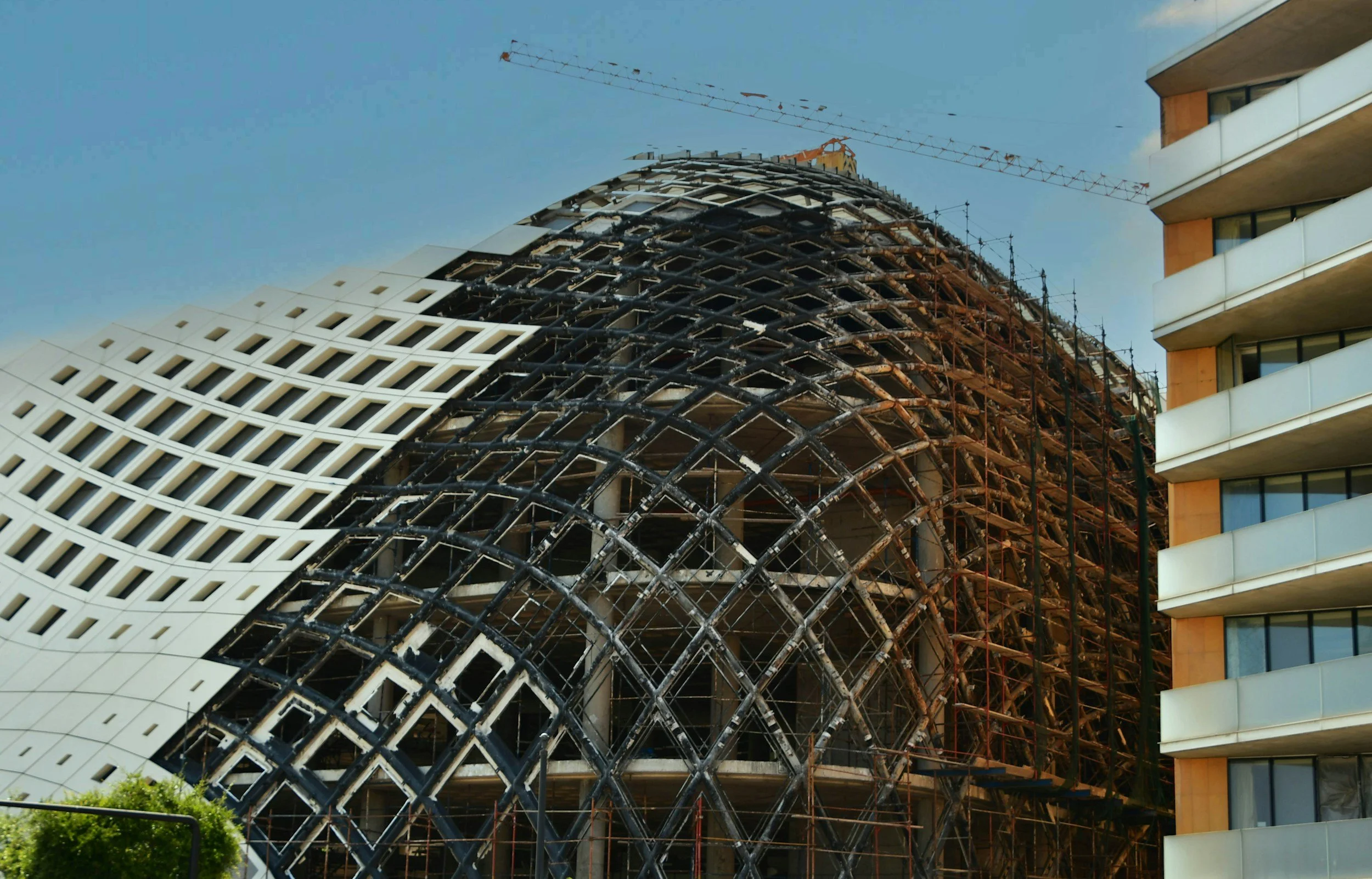 Bauarbeiten an einem modernen, gewölbten Gebäudeteil mit Gerüsten und Baumaschinen auf den Baustellen, während im Hintergrund ein Hochhaus sichtbar ist.
