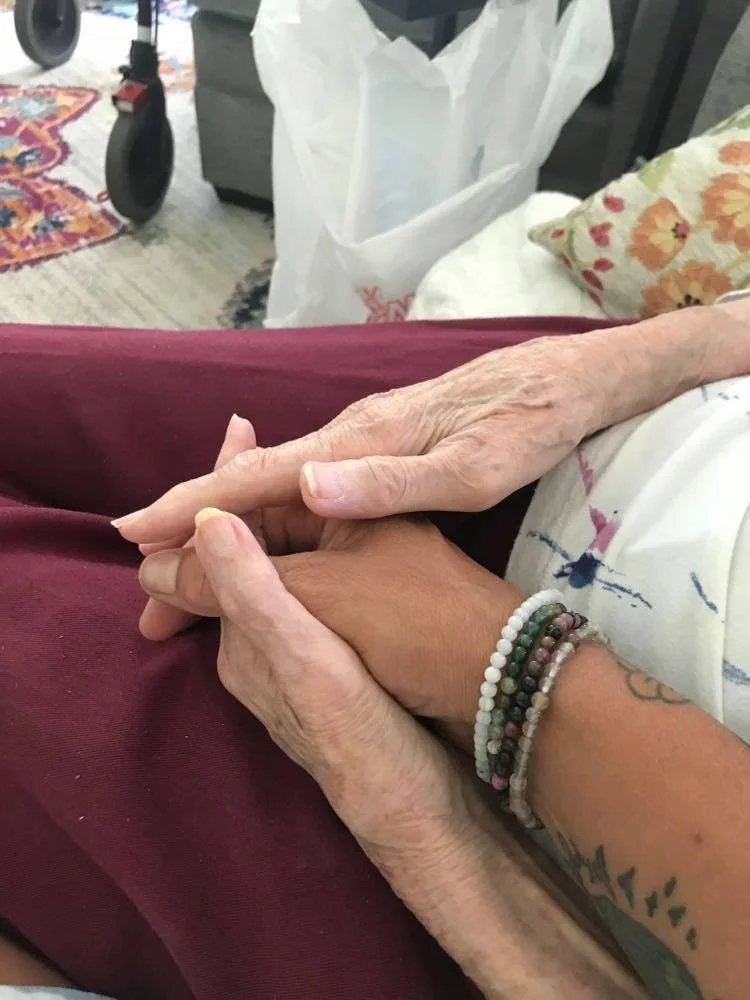 Close-up of two hands, one older and wrinkled, and one younger with a tattoo and bead bracelets, gently holding each other.