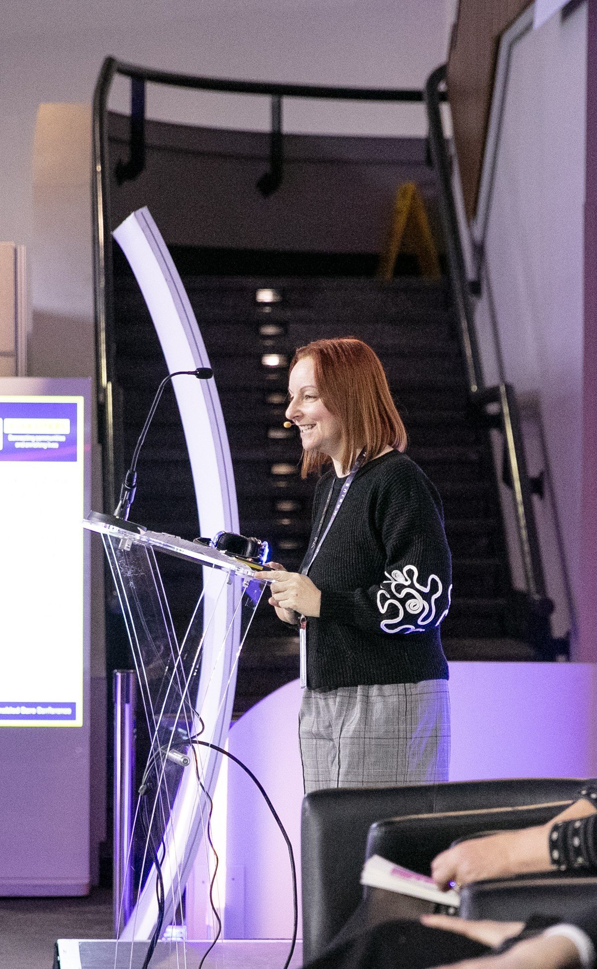 Jane Brightman is wearing a black sweater with a white design on the sleeve, stands at a transparent lectern with a microphone, smiling while speaking or presenting at an event or conference.