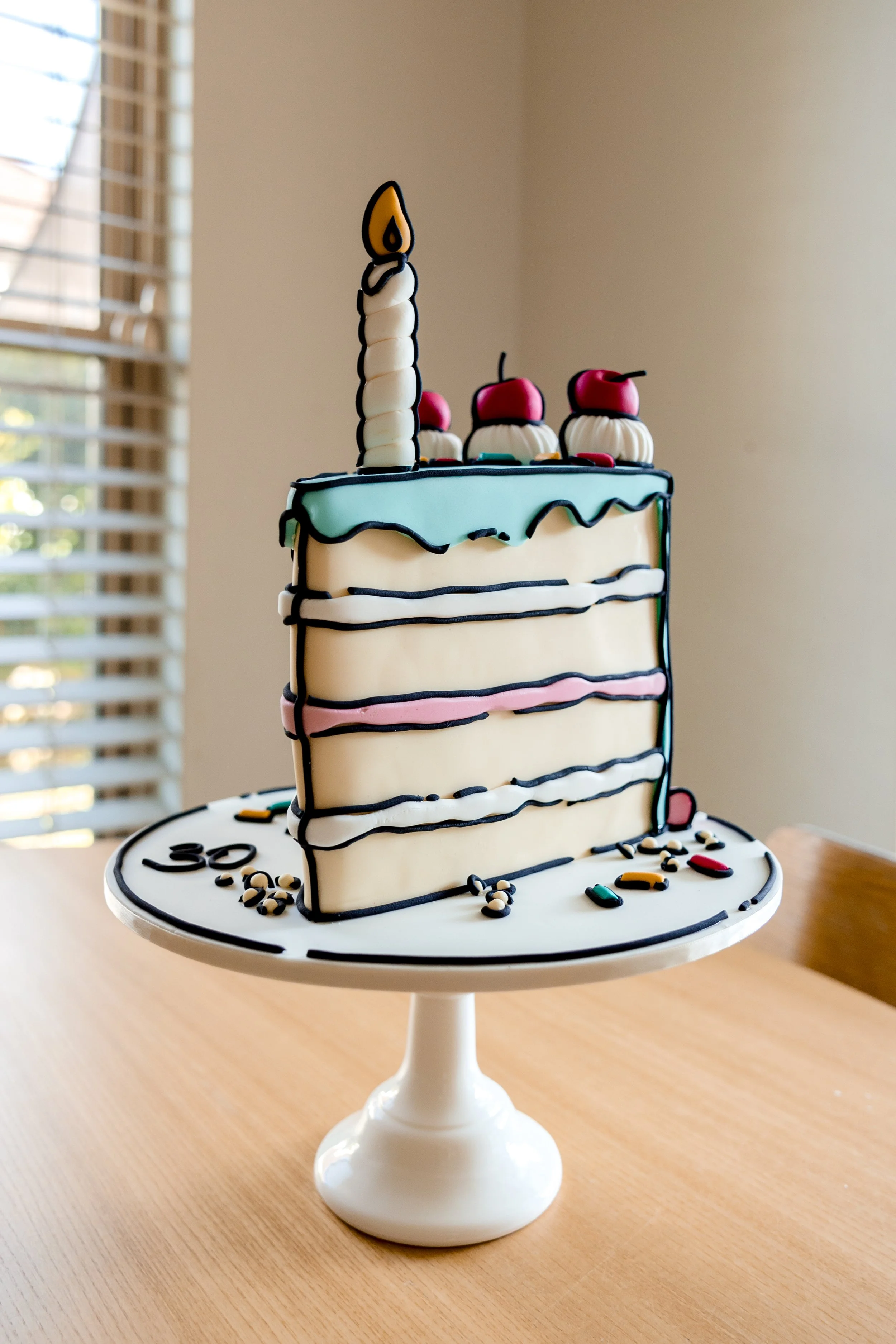 A birthday cake with white, pink, and blue icing, decorated with a single candle, cherries, and colorful candies on top. The cake is on a white cake stand on a wooden table.