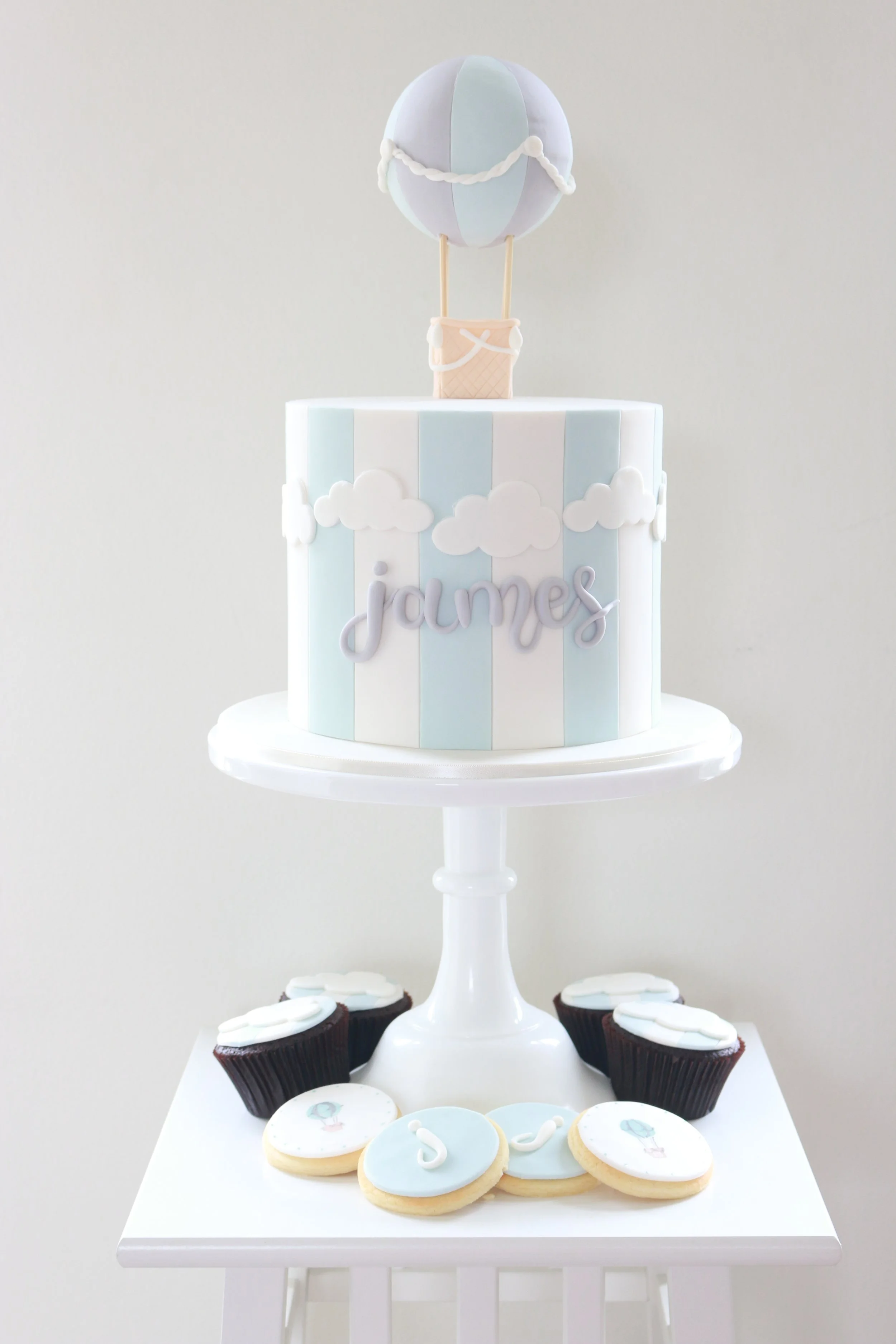 A decorated cake with pastel blue and white colors, featuring a hot air balloon topper, clouds, and the name 'james'; surrounded by cupcakes with similar themes on a white stand
