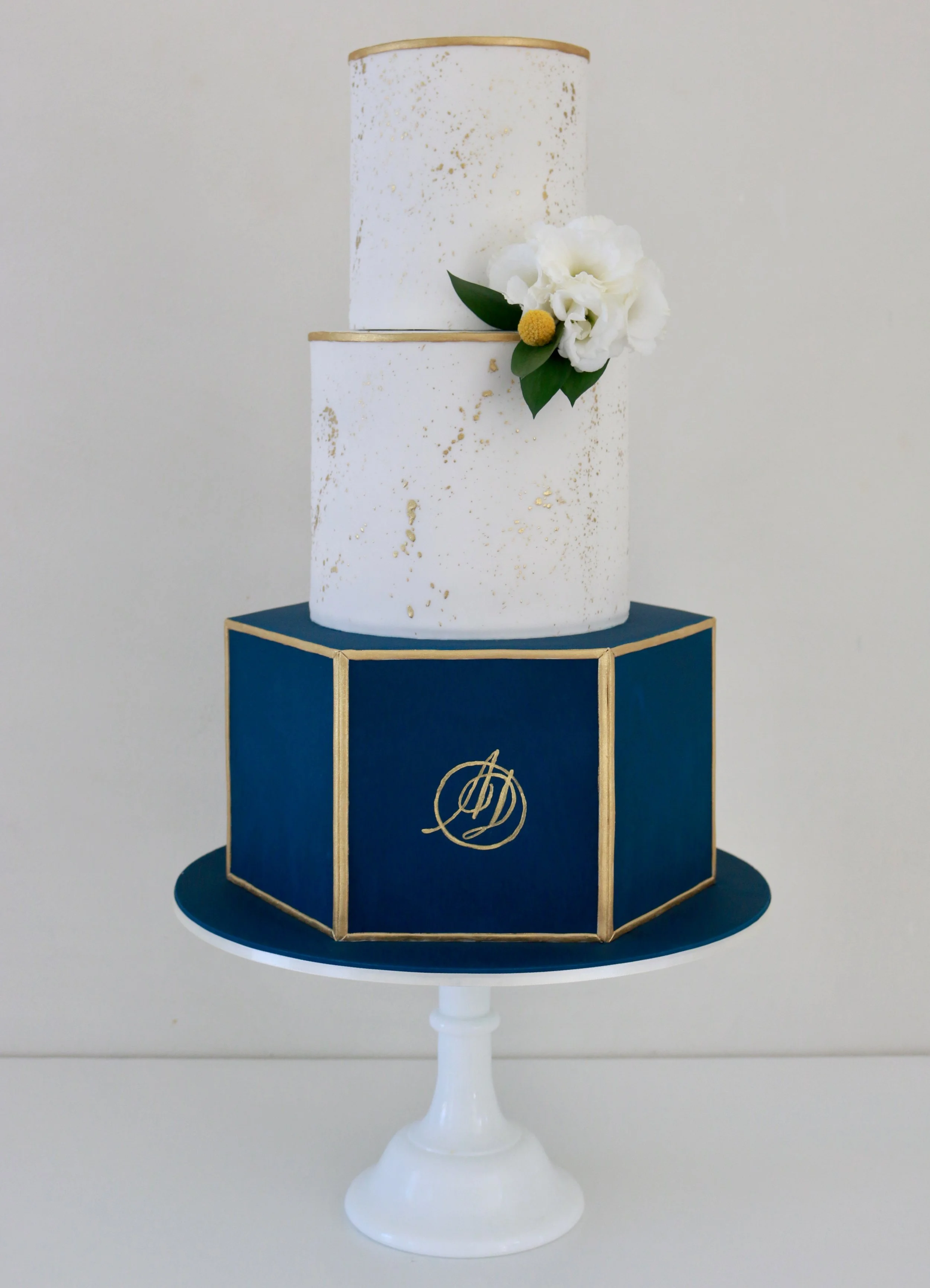 White two-tiered cake with gold specks and trim, decorated with white flowers and green leaves, sitting on a blue cake stand with gold accents, on a white table.
