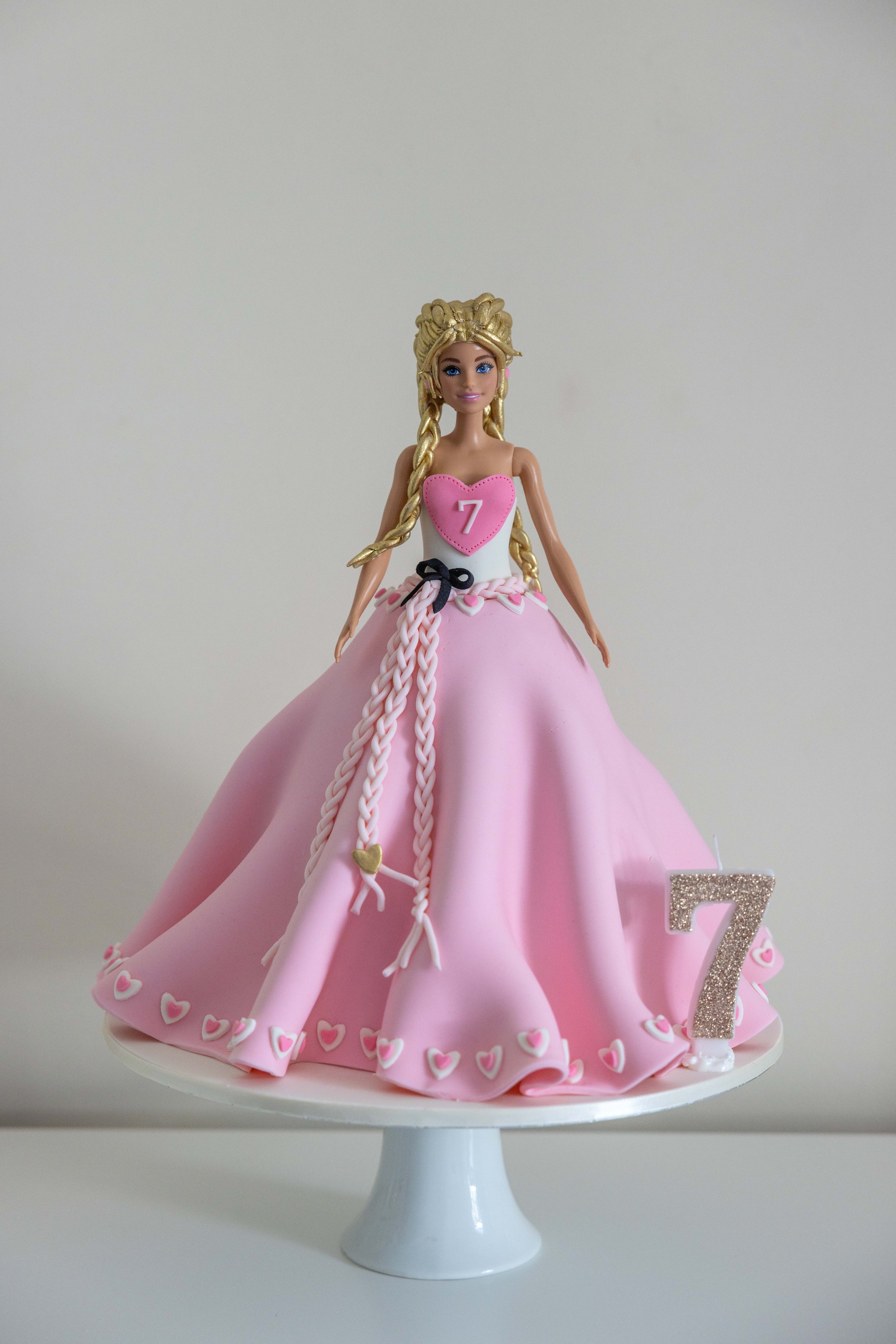 A Barbie doll cake decorated with pink and white accents, wearing a pink dress with a heart on the bodice and braids in her hair, displayed on a white cake stand with a glittery number 7 figure.