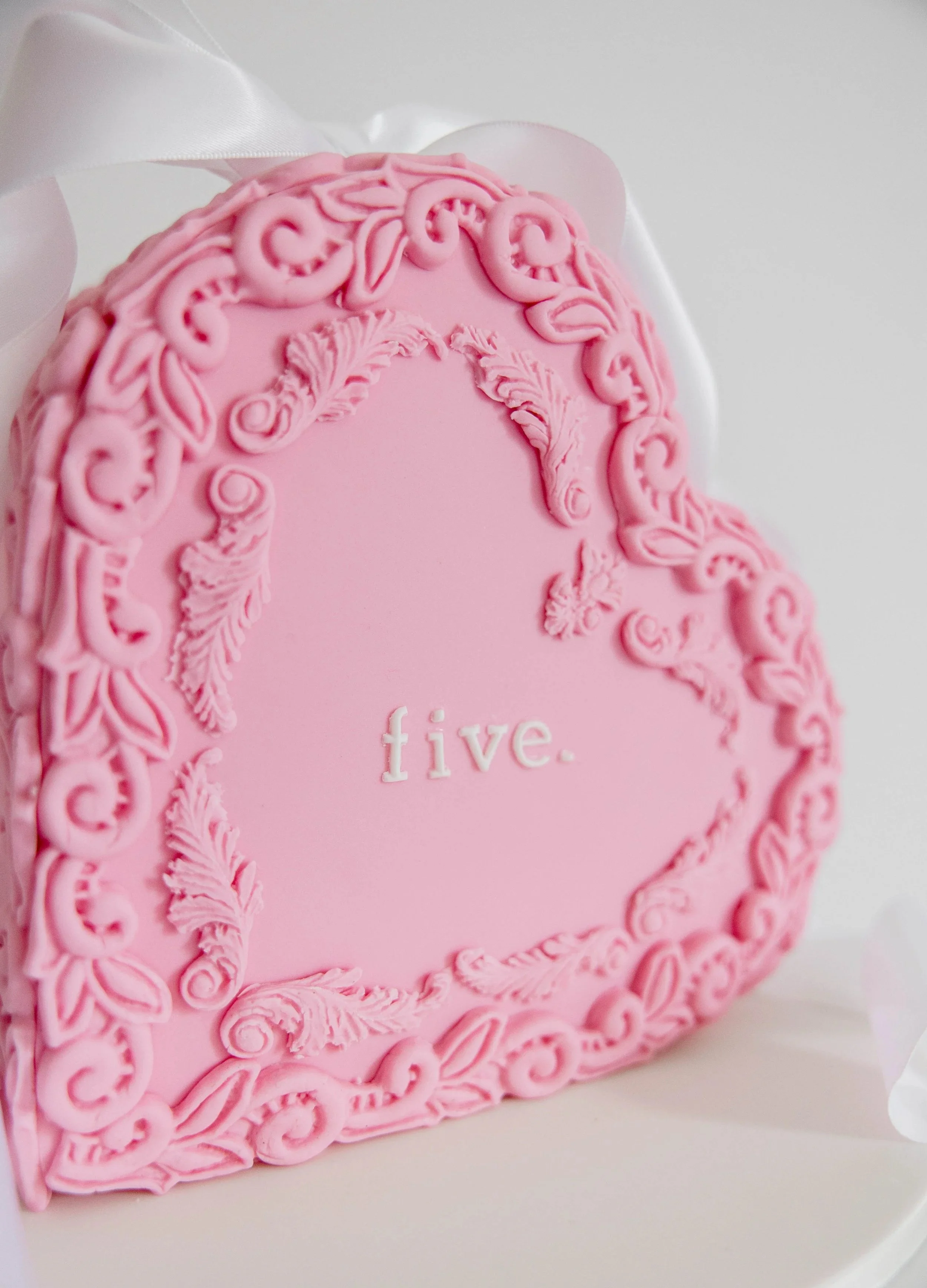 Close-up of a pink, decorative birthday cake with intricate icing designs and the word 'five' written on it, placed on a white plate with a satin ribbon in the background.