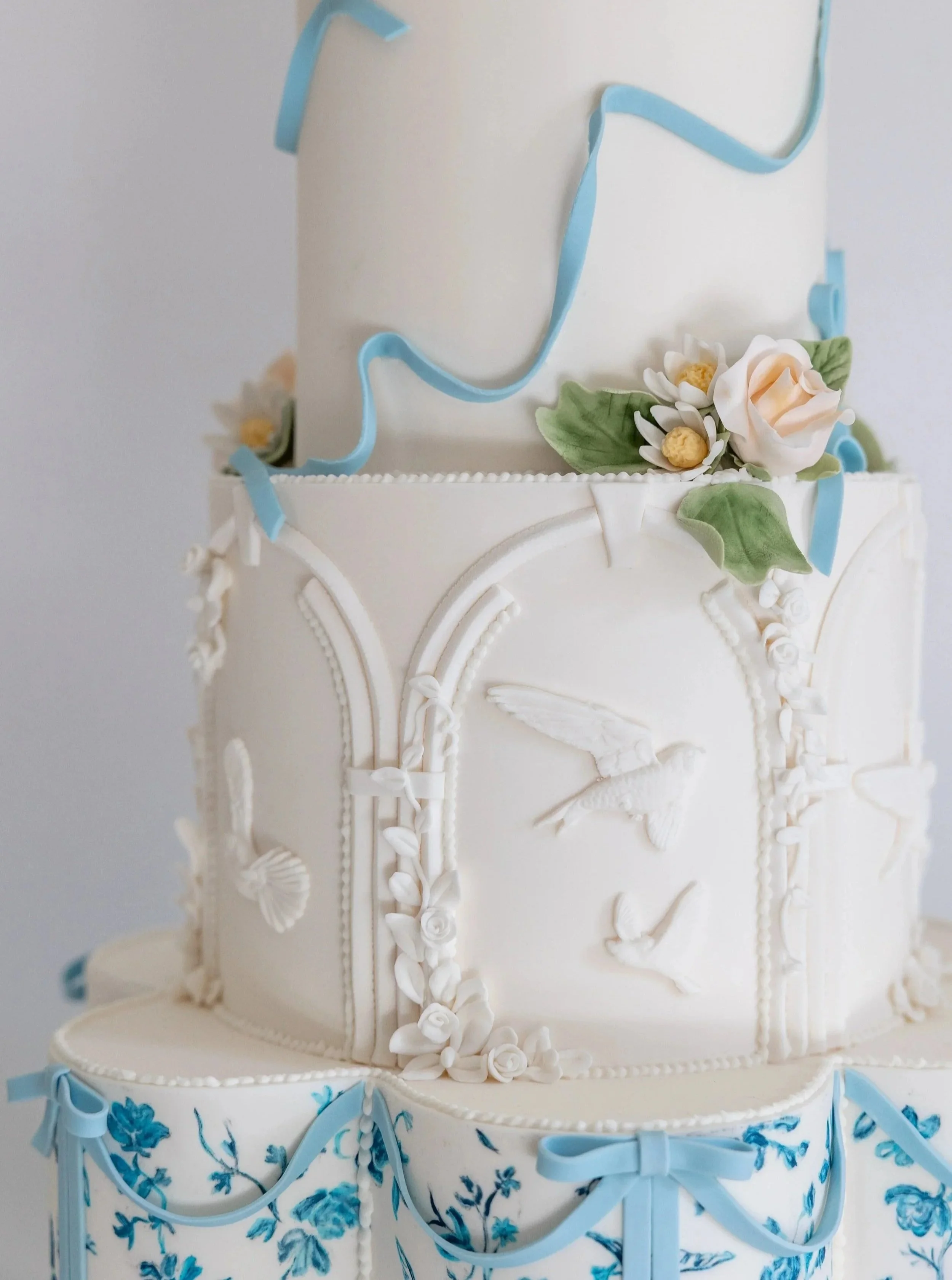 A white multi-tiered cake decorated with blue ribbons, white flowers, and intricate frosting details, with a blue floral pattern on the lower tier.