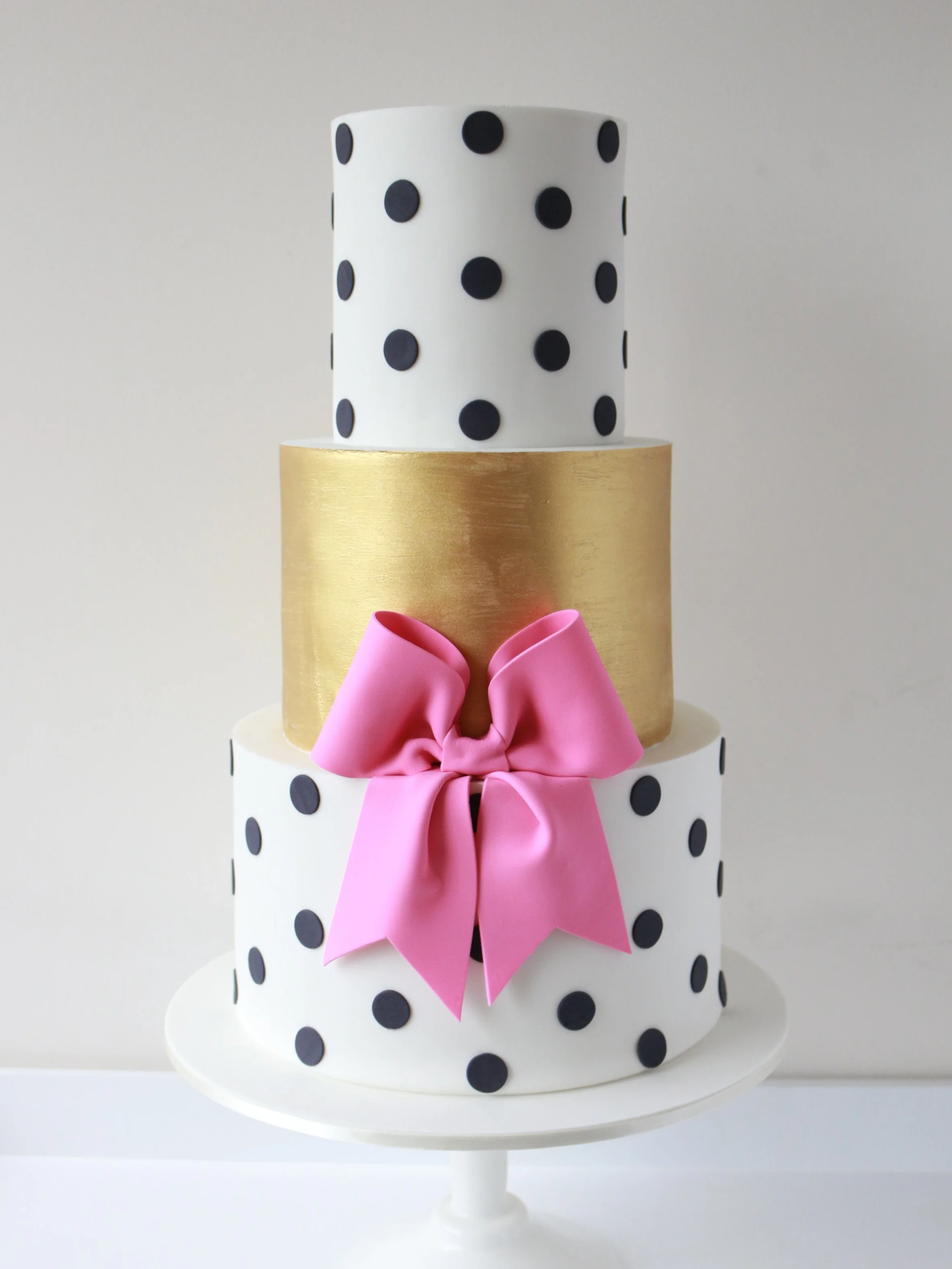 A tiered cake with black polka dots, a pink bow, a gold band in the middle, and a white base, set on a white cake stand against a plain background.