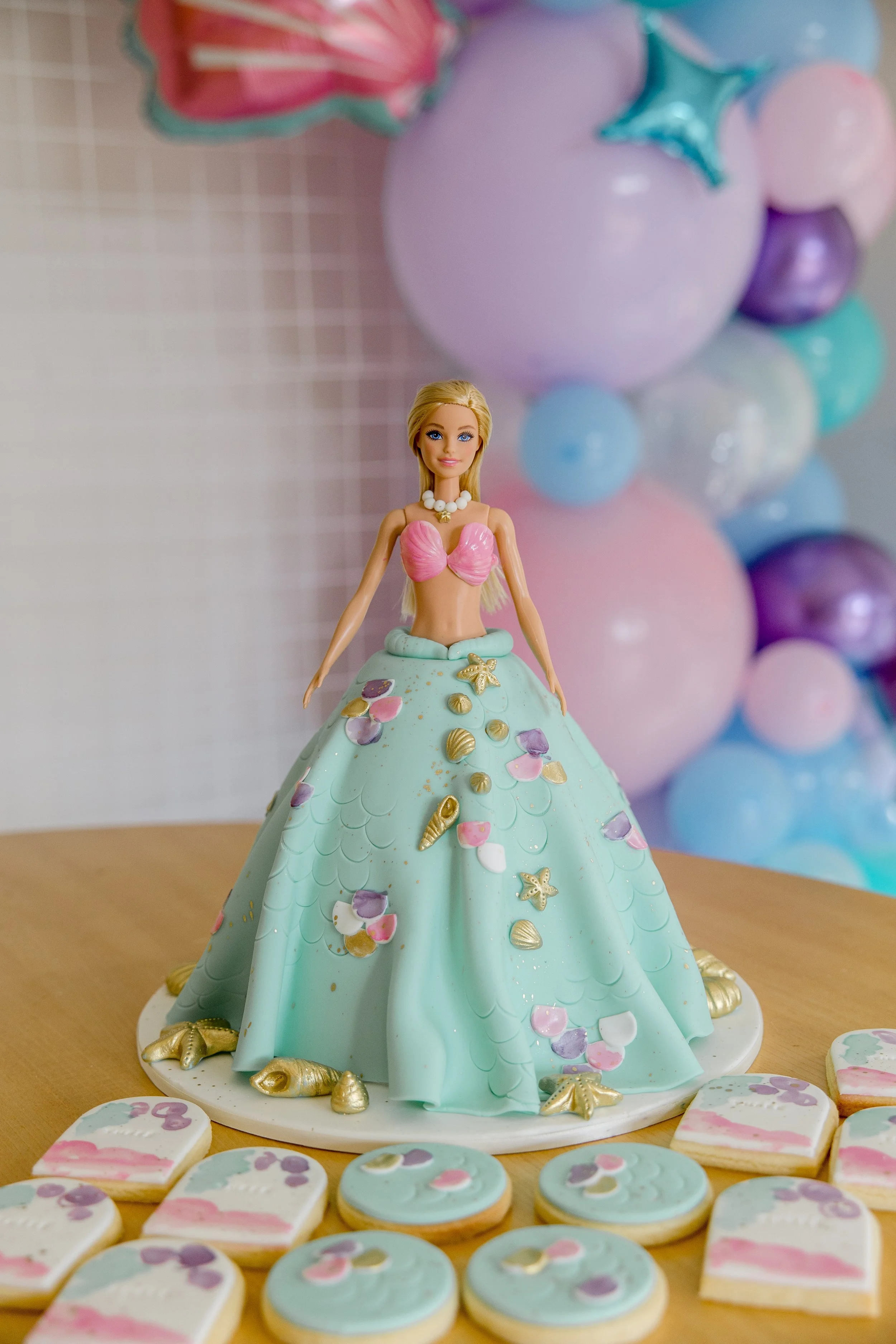 A cake shaped like a doll dressed in a pastel green ball gown decorated with seashells and starfish, with a doll figurine's head and torso on top, surrounded by pastel-colored balloons and decorated cookies on a wooden table.
