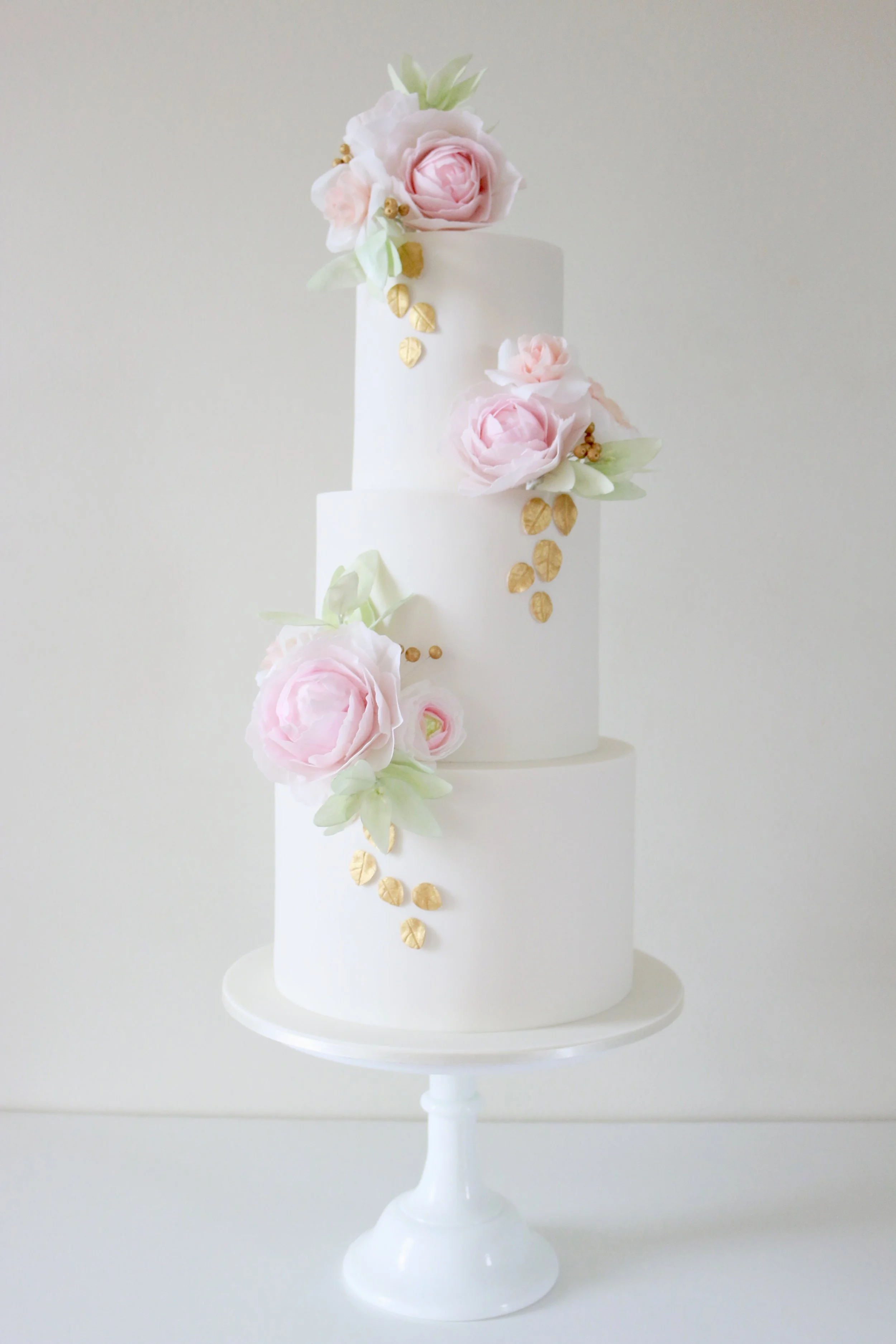 A three-tier white cake decorated with pink roses, white flowers, green leaves, and gold-colored leaves, displayed on a white cake stand against a neutral background.