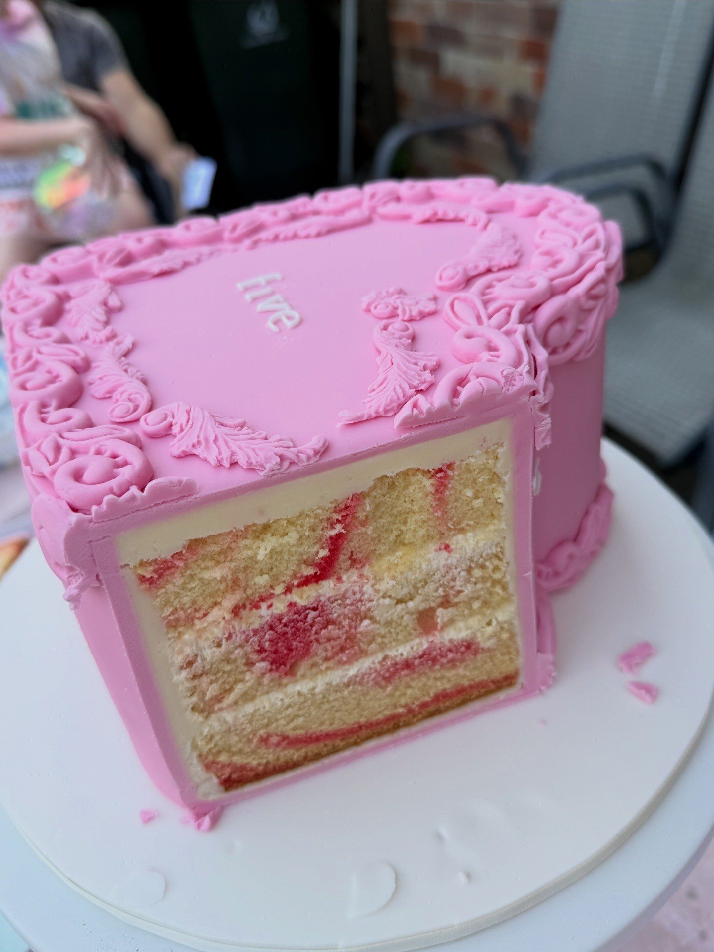 Close-up of a pink, heart-shaped layered cake with pink icing decoration and the word 'love' written on top, with a slice removed to reveal the cake's interior layers.