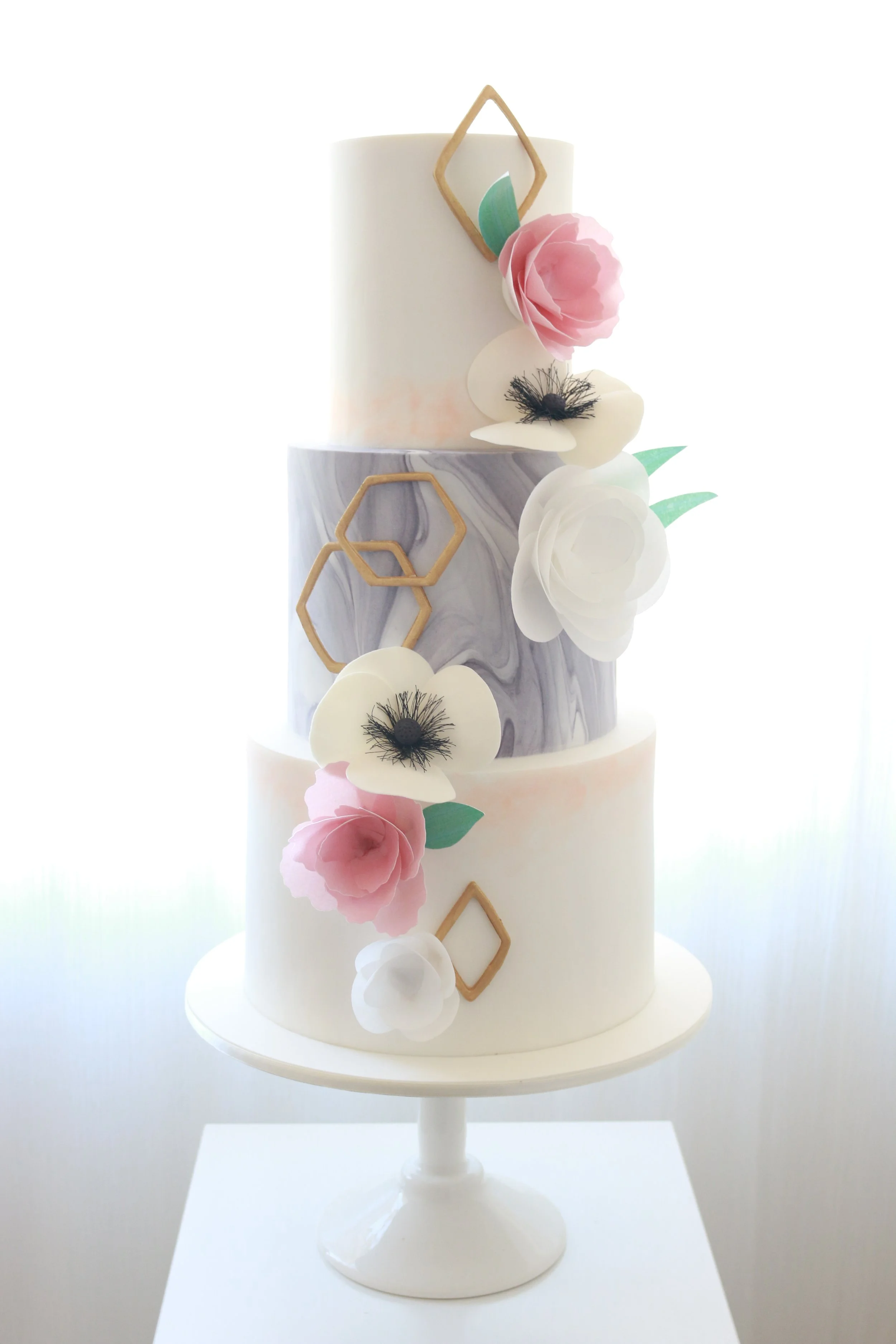 Two-tier white wedding cake decorated with pink and white flowers, green leaves, and gold geometric accents.