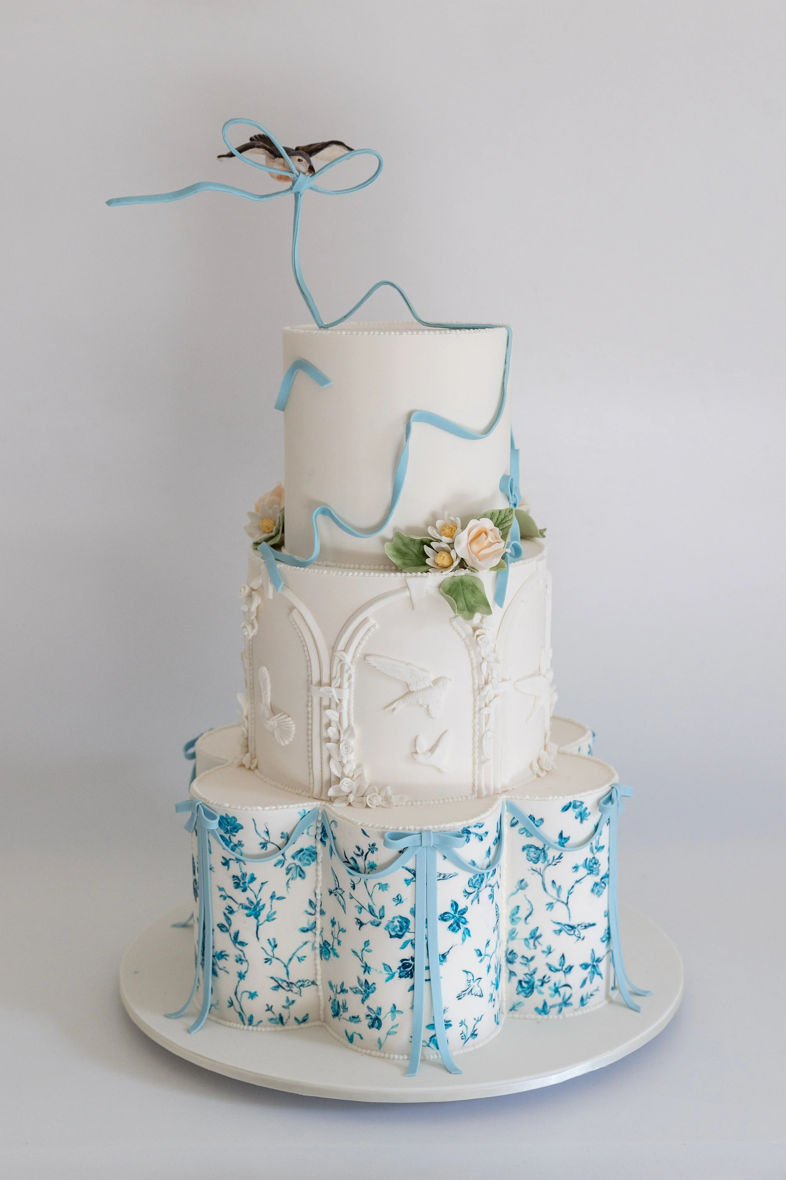 Three-tiered white wedding cake decorated with blue ribbons and floral patterns, topped with a wire sculpture of a bird.