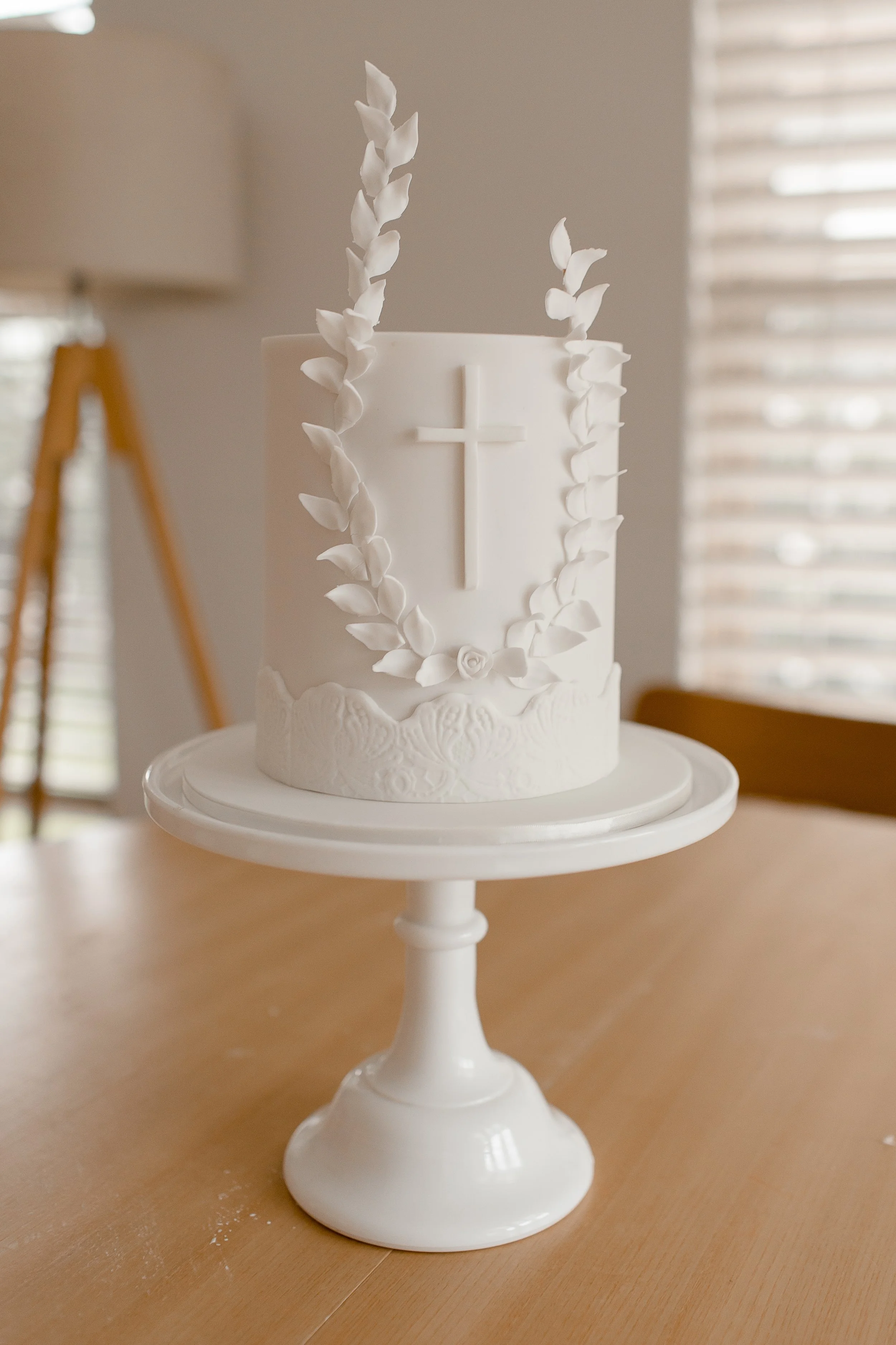 White wedding cake with embossed pattern, adorned with a cross and leafy decorations, placed on a white cake stand in a room with window blinds.