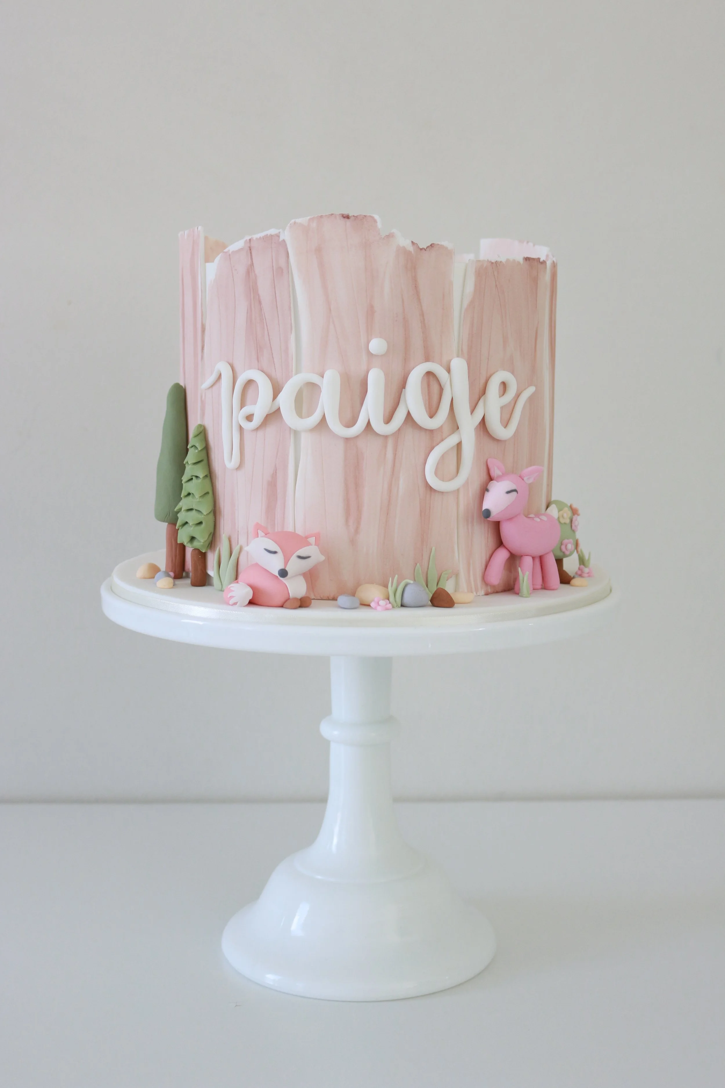 A cake with pink and white marbled icing, decorated with fondant animals including a fox and a deer, trees, rocks, and the message 'I love papa' in white cursive letters, sitting on a white cake stand.